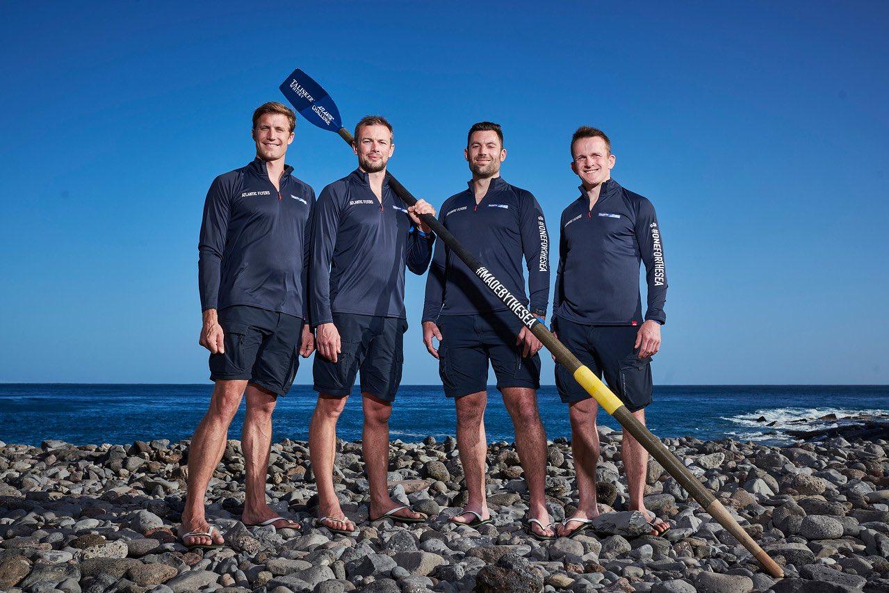 Rowers stand on pebble beach with sailing oar.