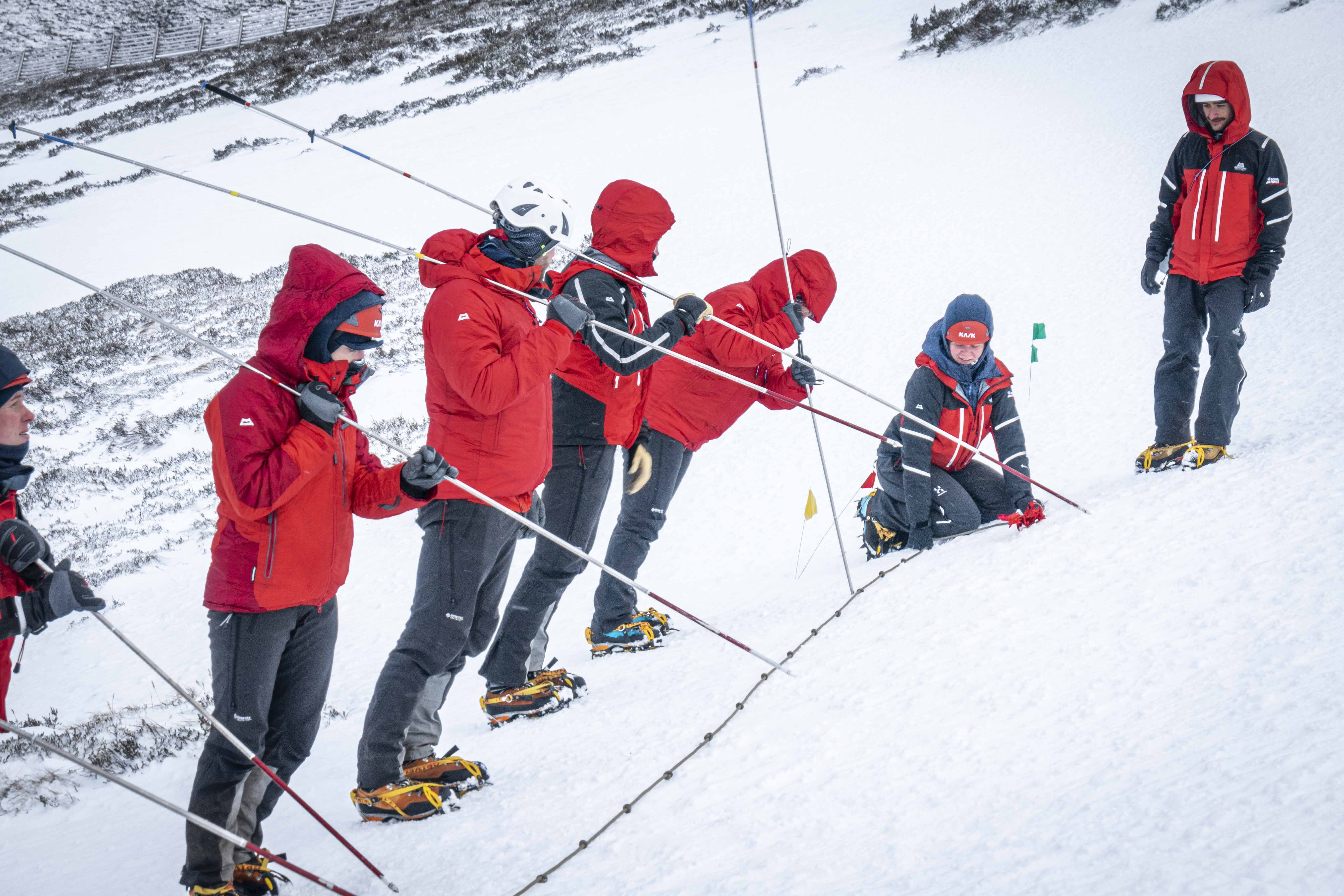 Image shows Mountain Rescue Team on a snowy mountain holding long poles and climbing equipment.
