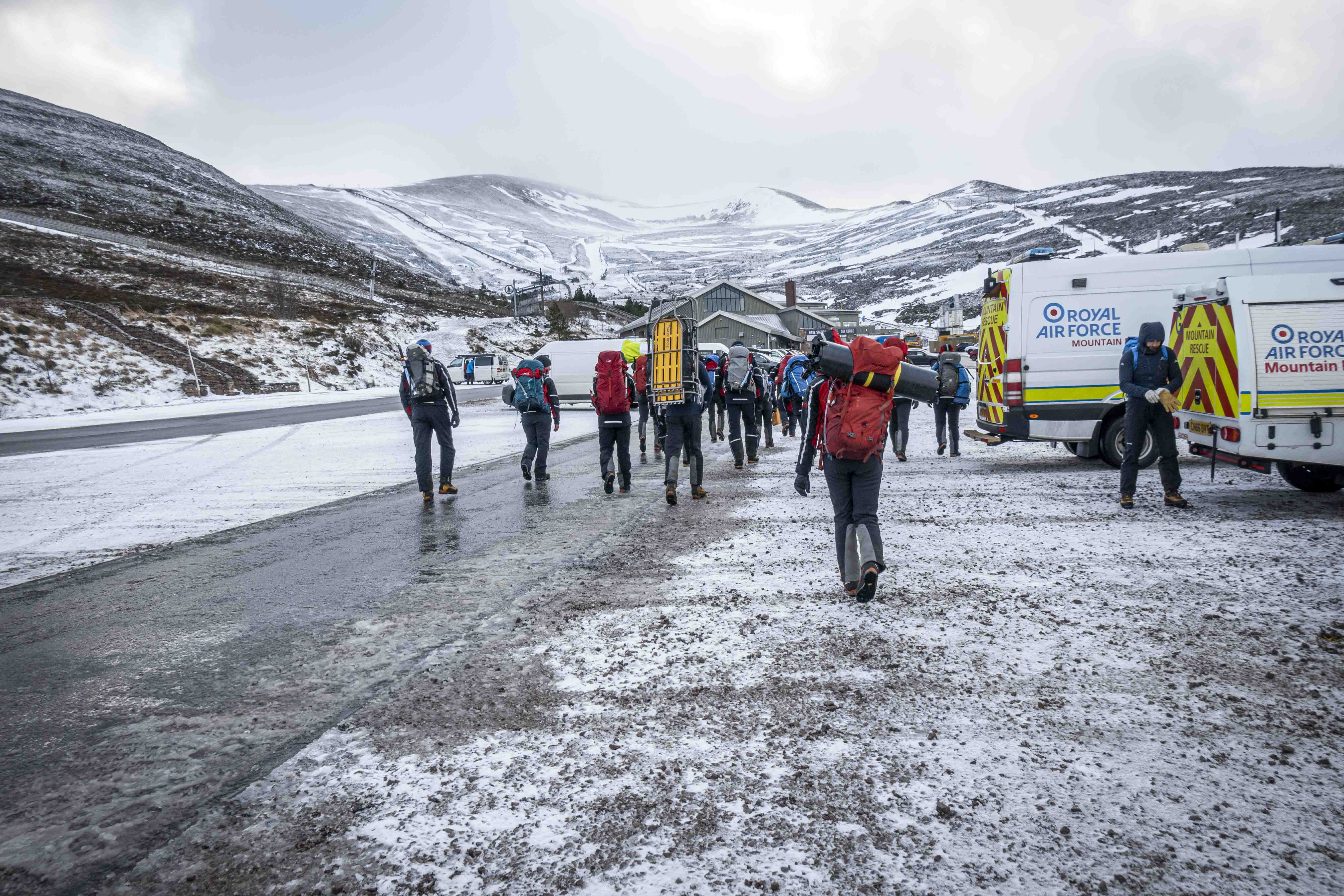 Image shows Mountain Rescue Team walking on a flat area between snowy mountains and vehicles.