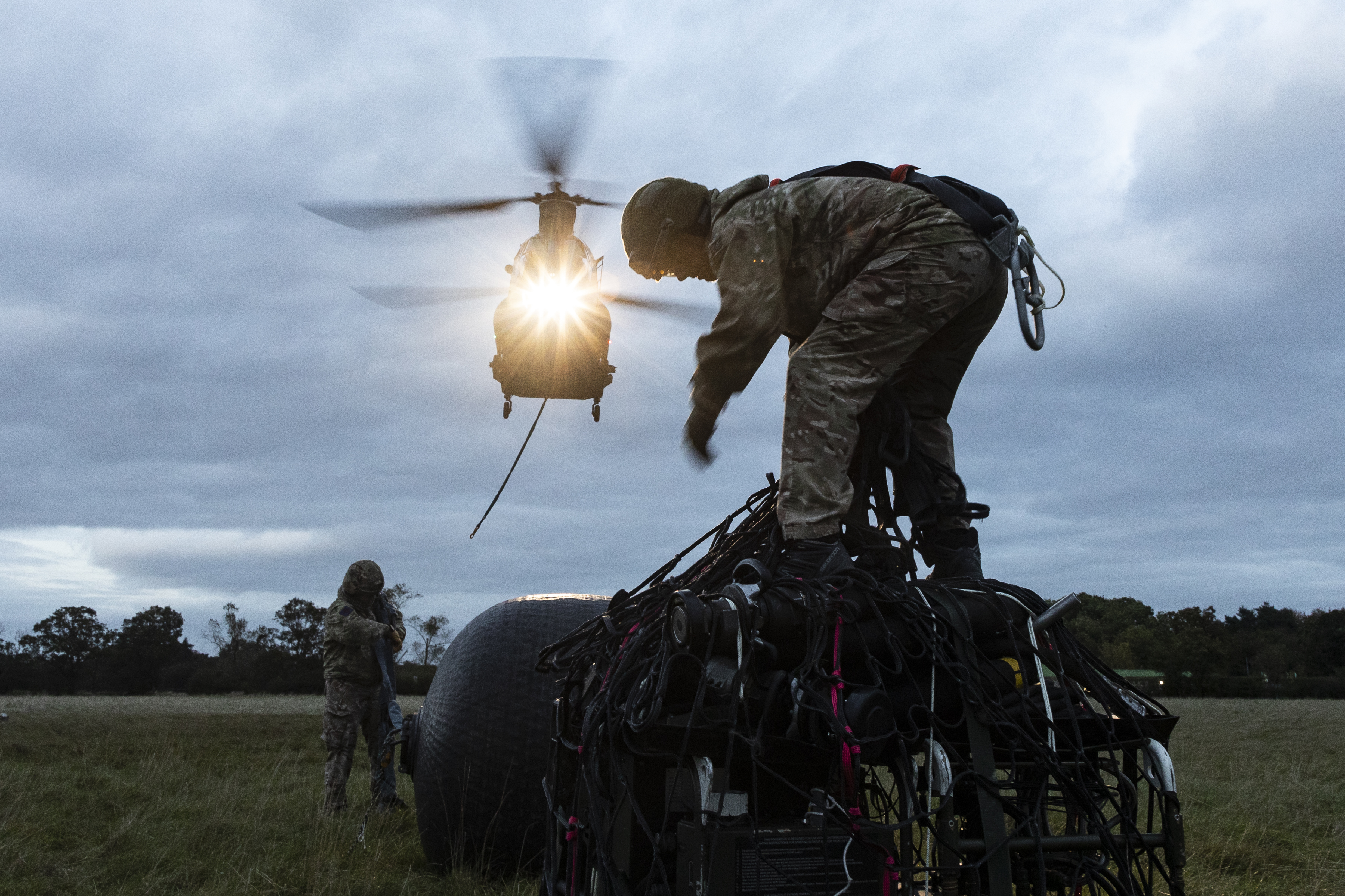A soldier from 23 Parachute Engineer Regiment stands on top of a netted load ready to hook it under the approaching Chinook, which can be seen with its lights on as it approaches from the ground