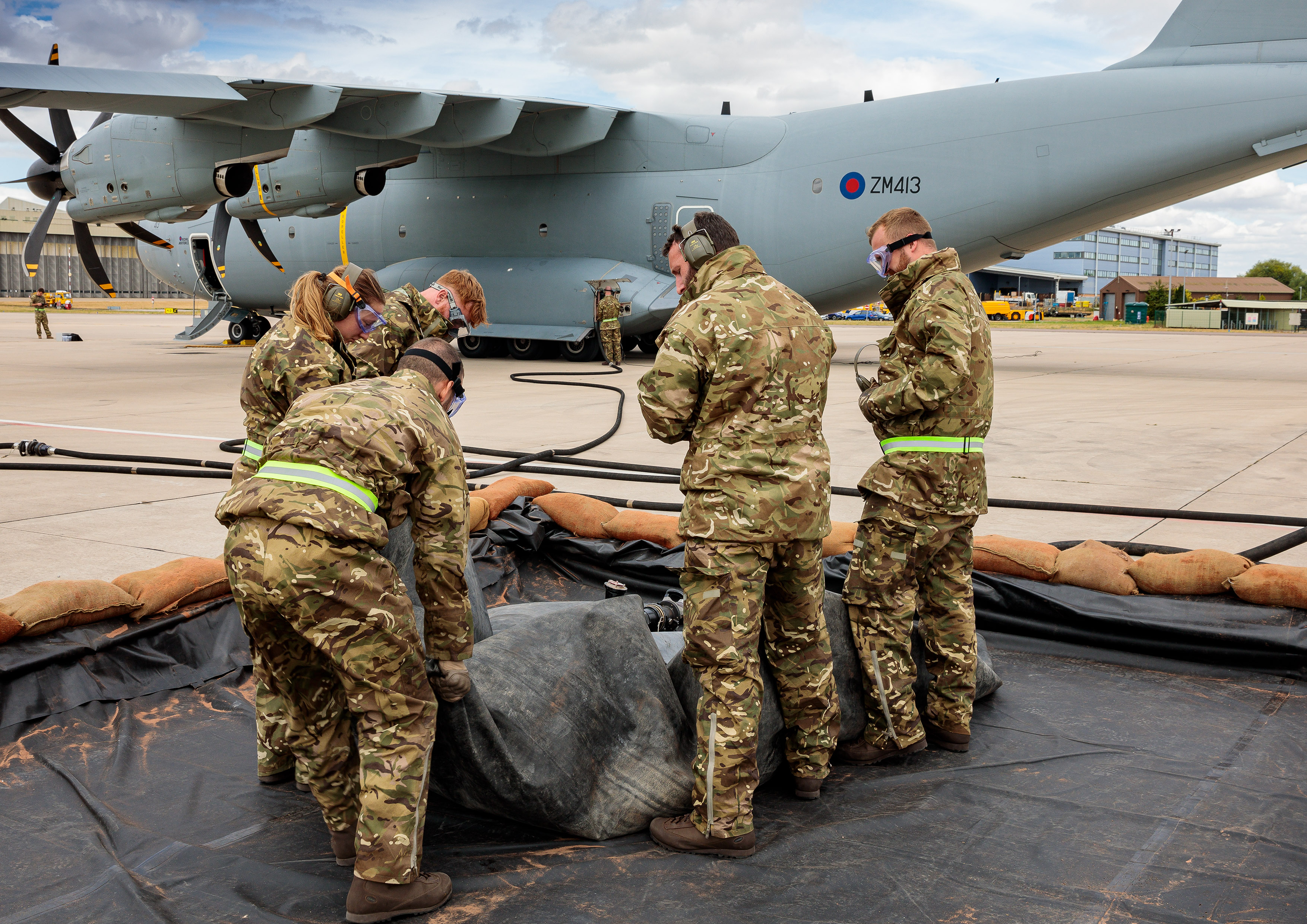 Image shows RAF aviators moving tarpaulin with Hercules aircraft in background. 