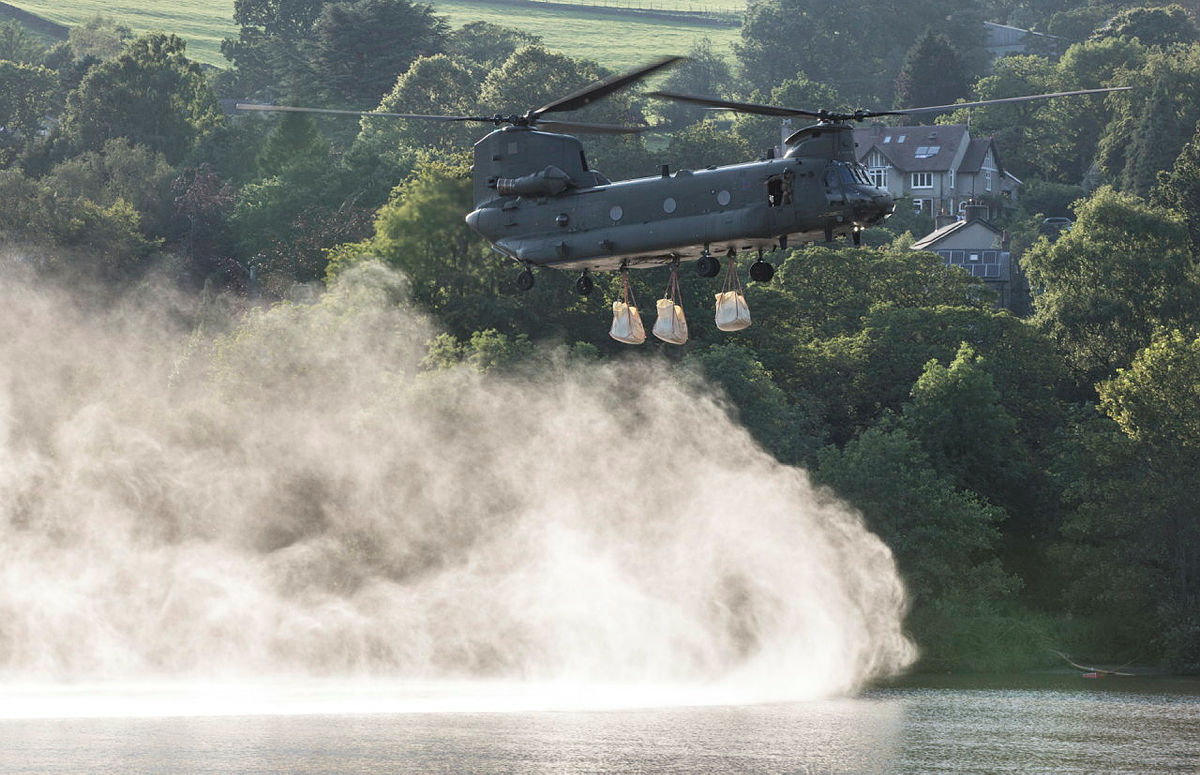 Image shows a Chinook flying low with underslung load, over lake. 