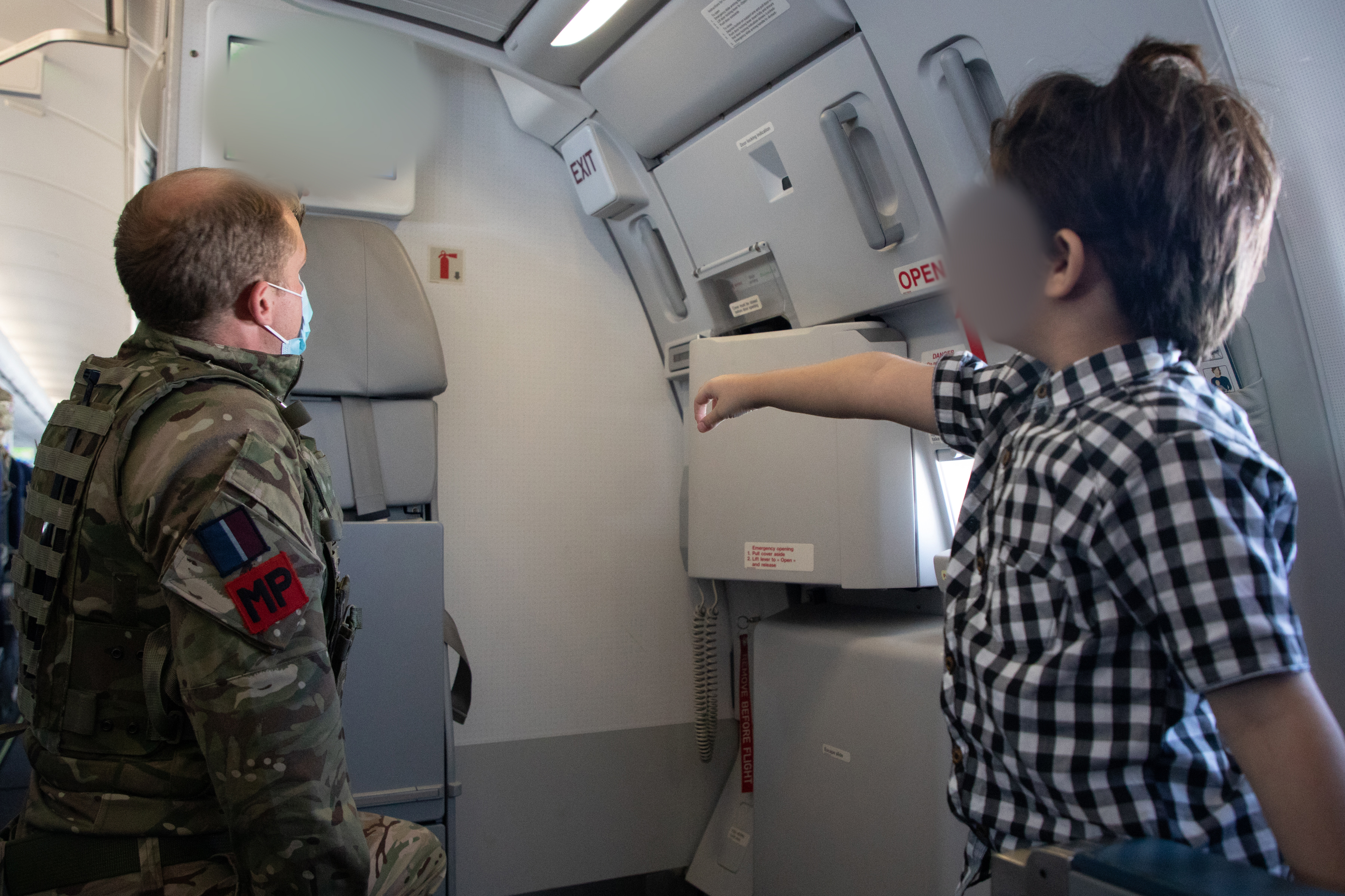 RAF Police and young passenger look at emergency escape door inside aircraft.