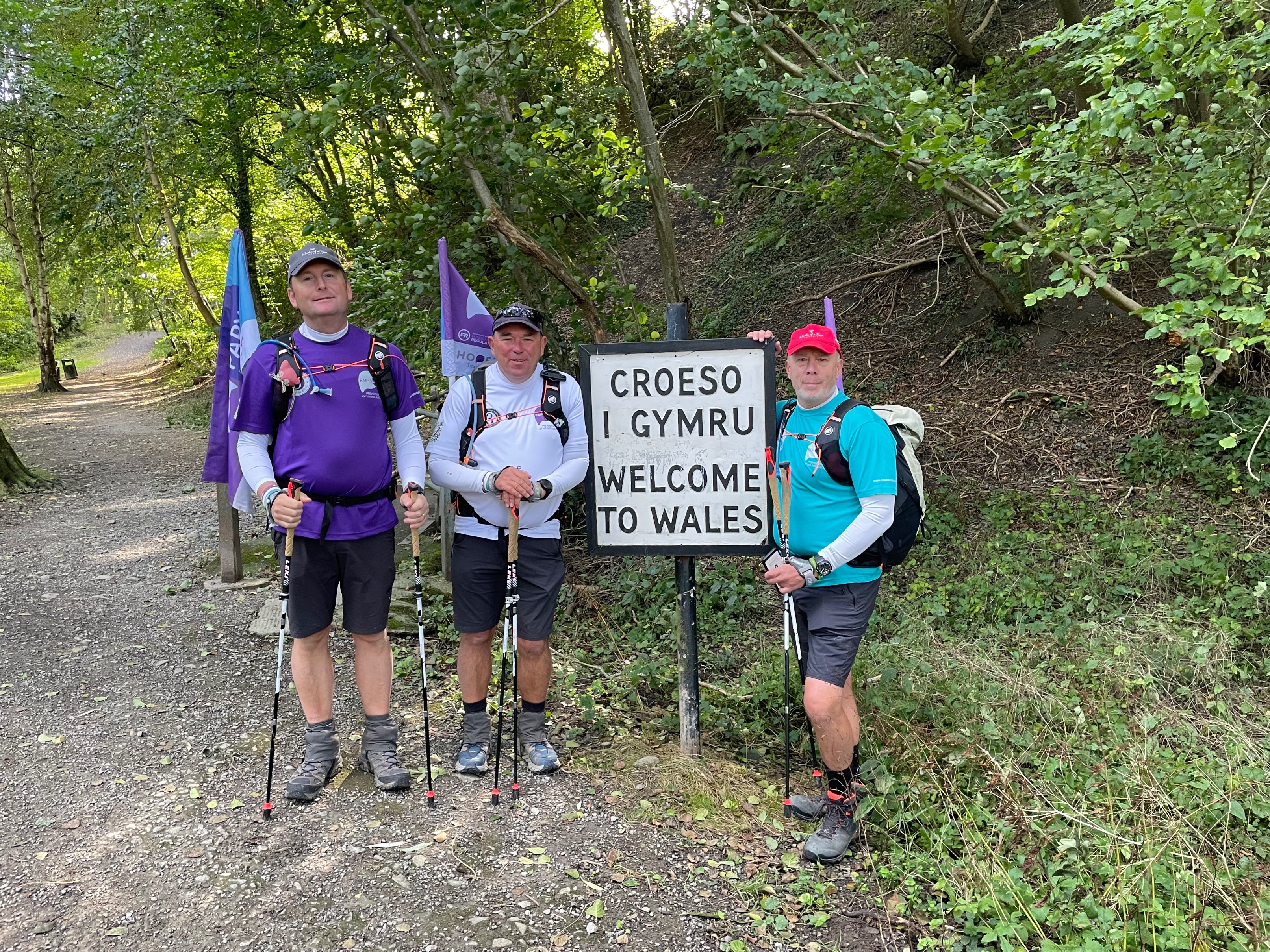 Image shows three dads by a welsh sign as they hike.