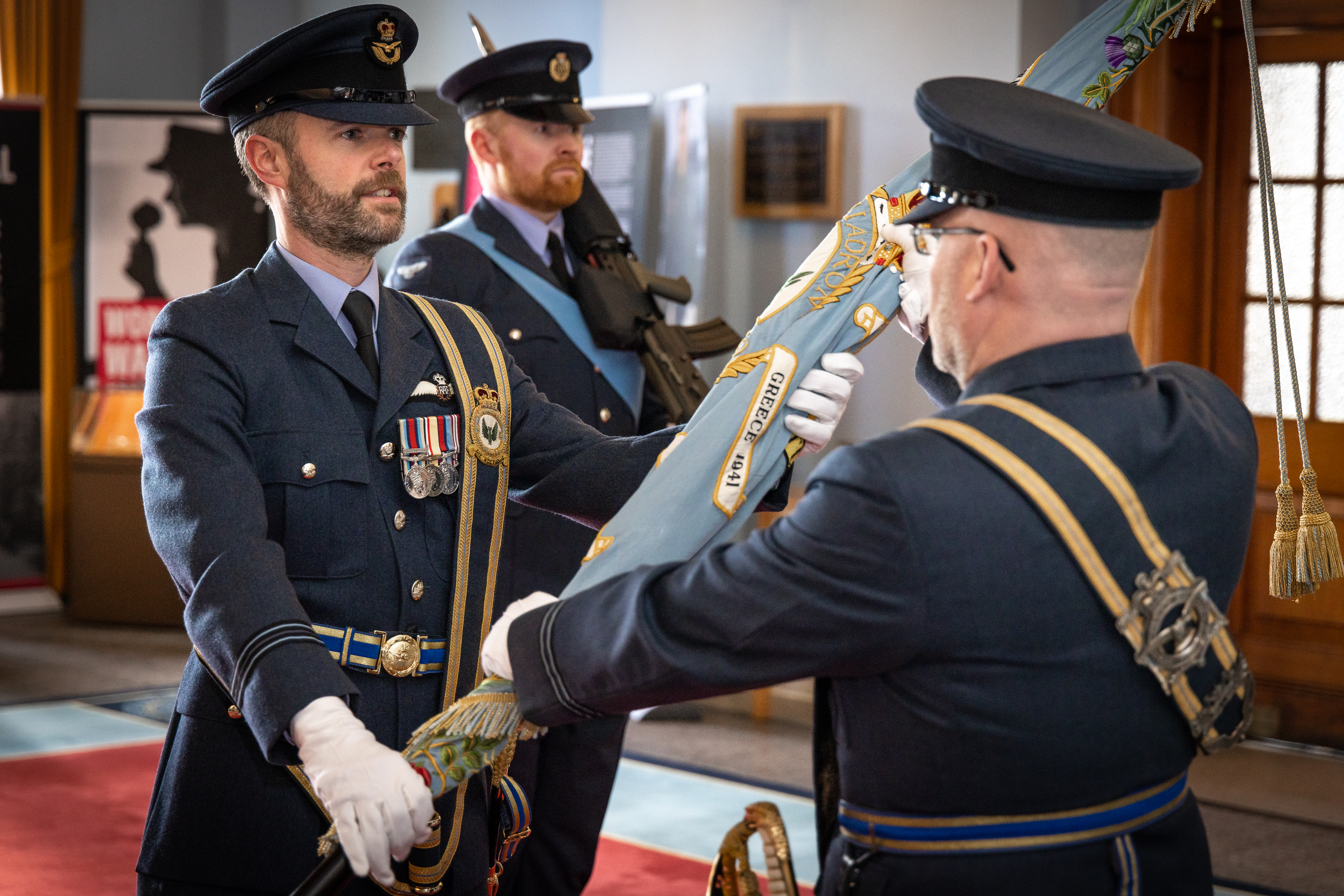 Image shows RAF aviators holding colour standard inside Officers' Mess during ceremony.