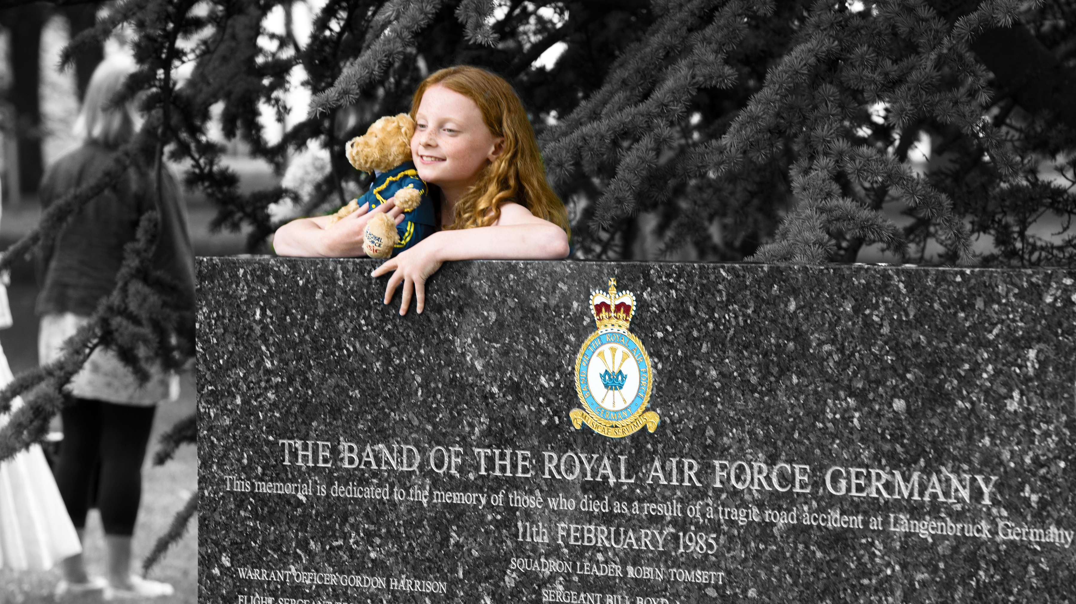 Girl and teddy bear leans on memorial.
