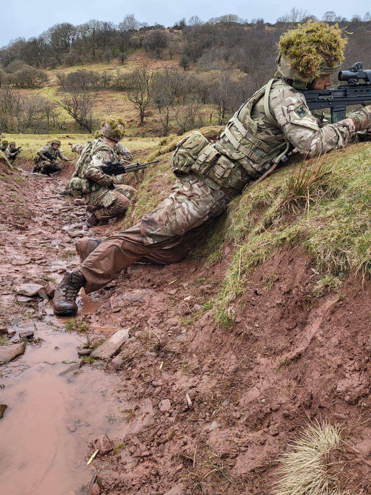 Personnel crouch in muddy river trench, with rifles.