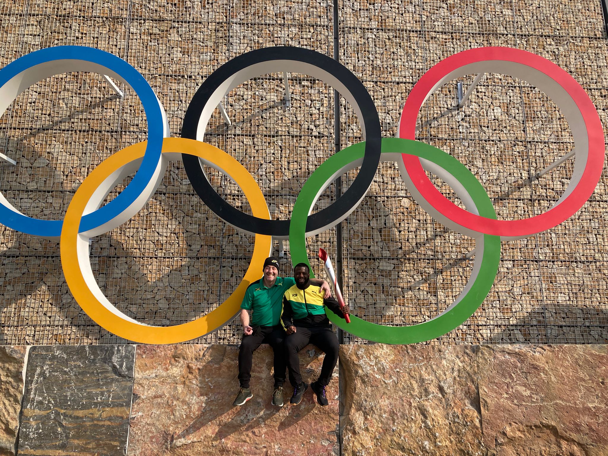 Shanywayne holds Olympic torch with a friend, while sitting on rocky ledge by Olympic rings.
