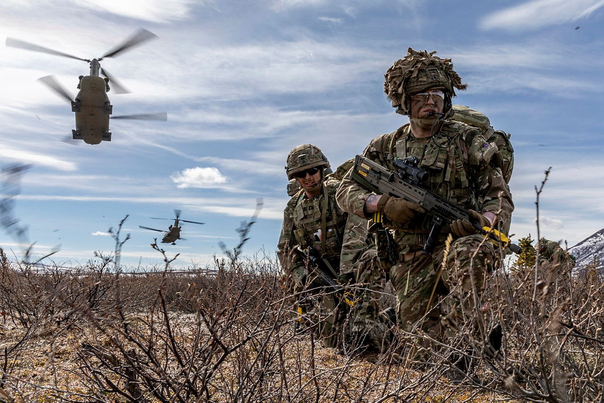 Image shows RAF Regiment patrolling with rifles, as a helicopter flies overhead.