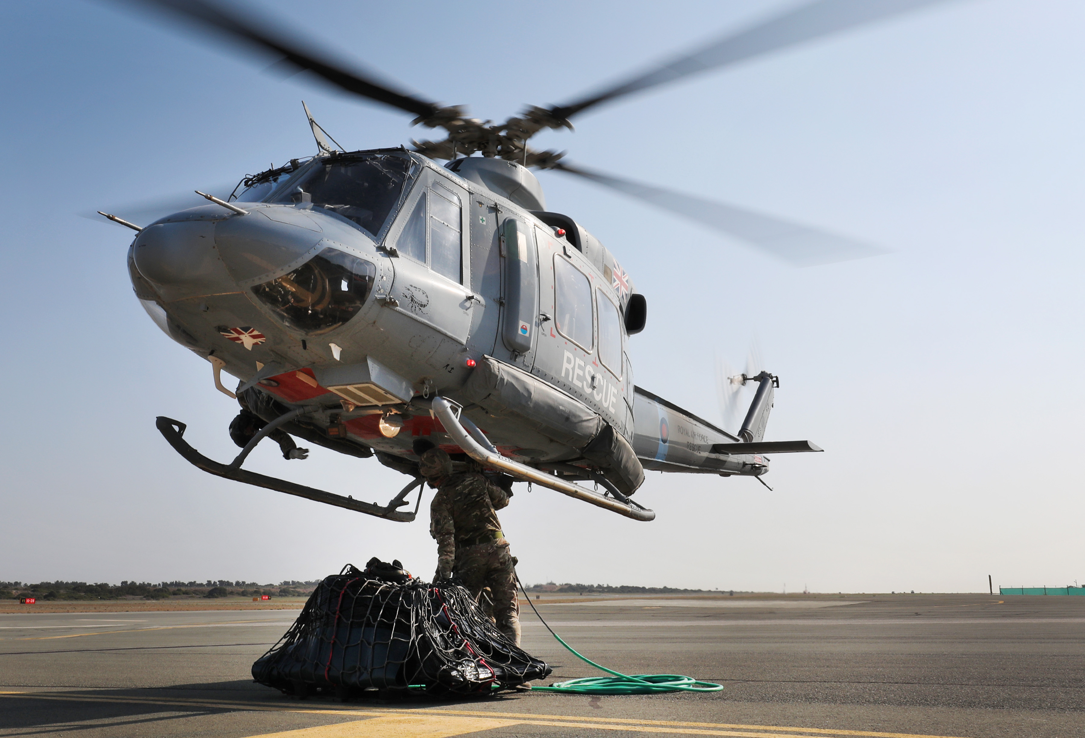 Image shows RAF aviators attaching cargo to the under slung load carrier of a Chinook helicopter, as it hovers overhead.