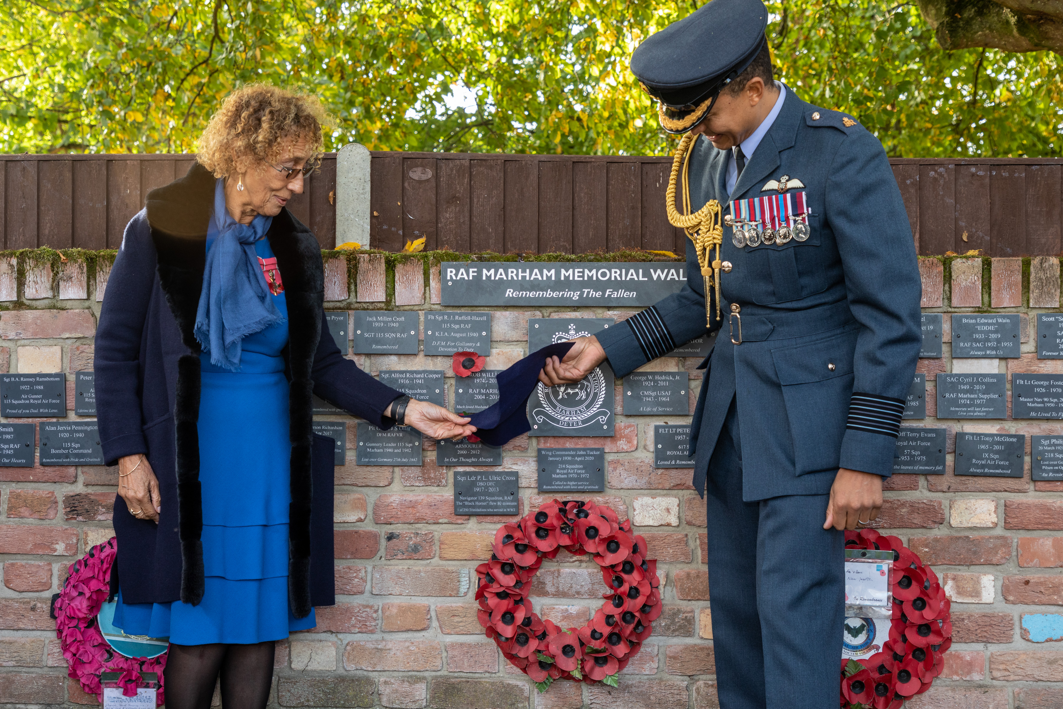 Image shows RAF aviator and civilian removing a cloth from a small plague on a memorial wall.