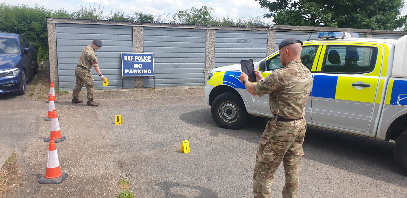 Image shows RAF Police taking pictures and setting out cones to mark a scenario crime scene in a garage car park, with their emergency vehicles.