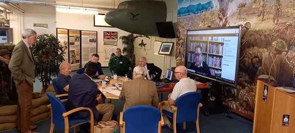 Image shows retired personnel around a table with a television and nose of carrier aircraft trophy mounted on the wall.