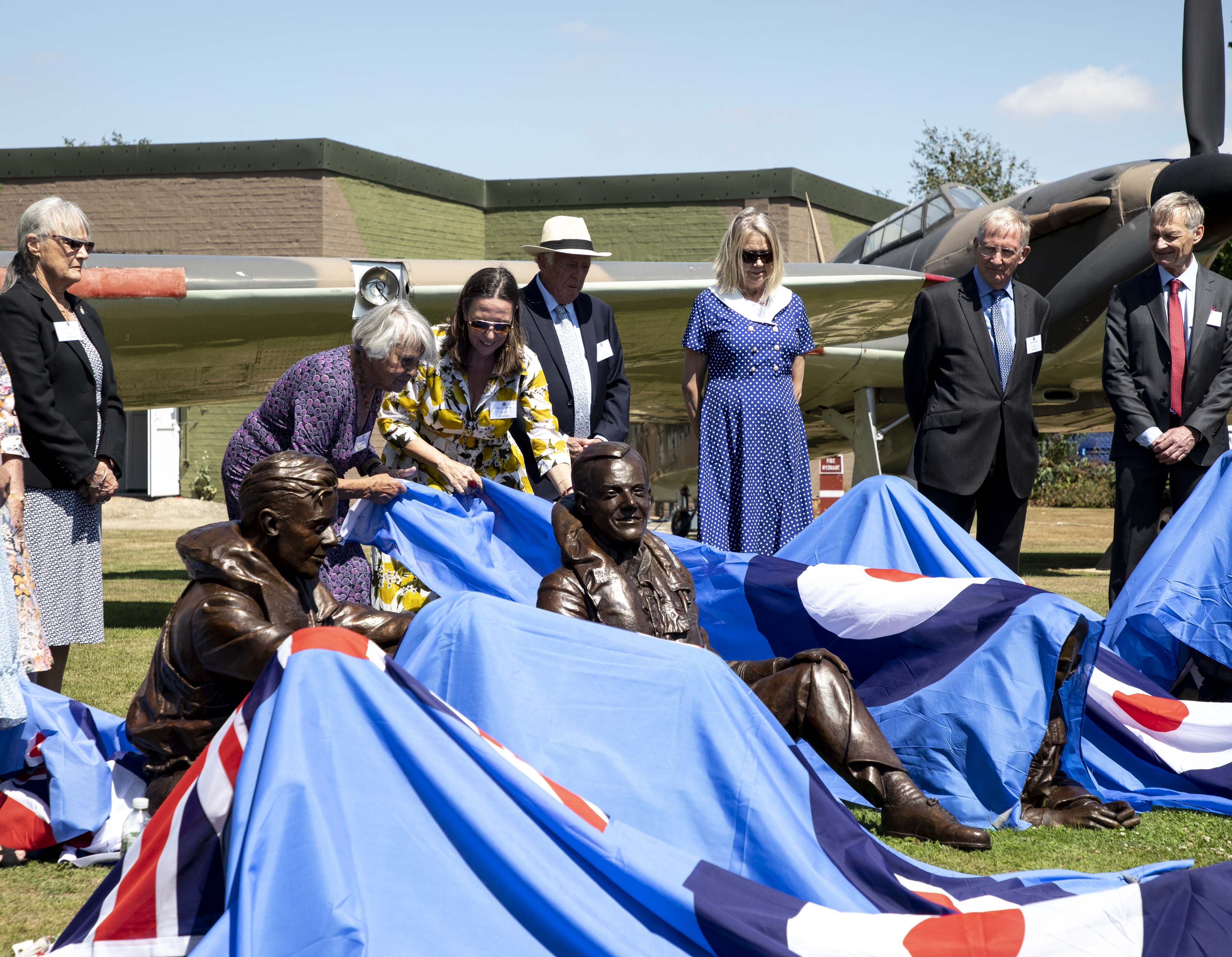 Image shows aviators and members of the public with the bronze sculptures from under blue coverings; with Hurricane aircraft in the background.