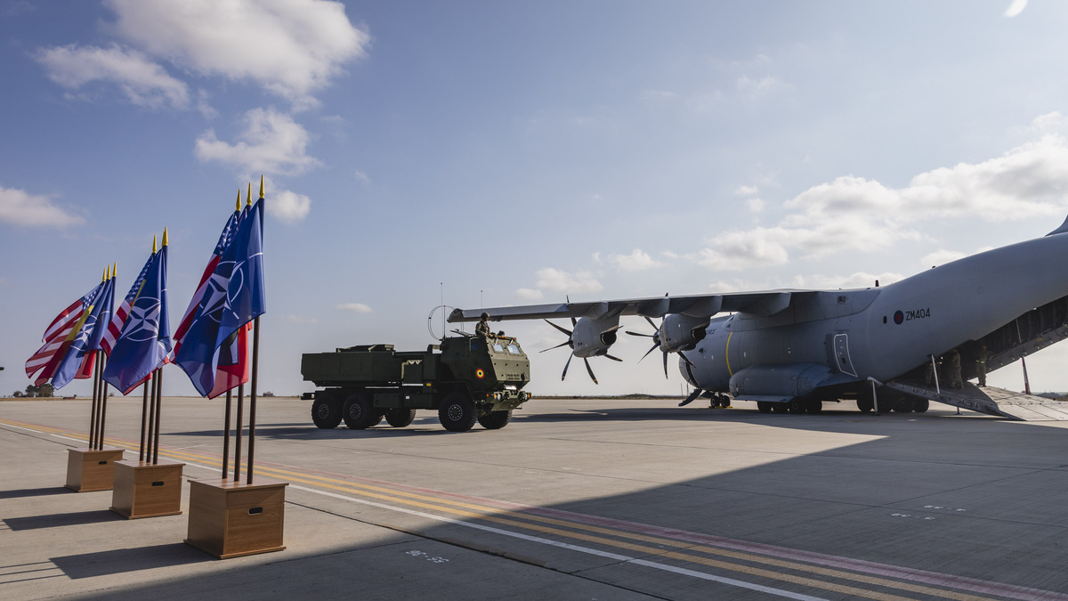 Image shows flag stand, truck, and Atlas aircraft with its loading bay open on an airfield.