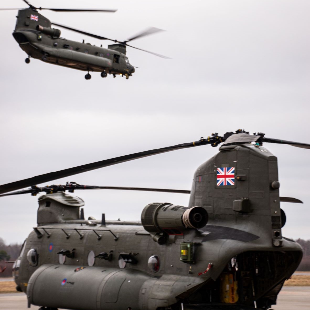 Image shows RAF Chinook flying over another Chinook on the airfield.
