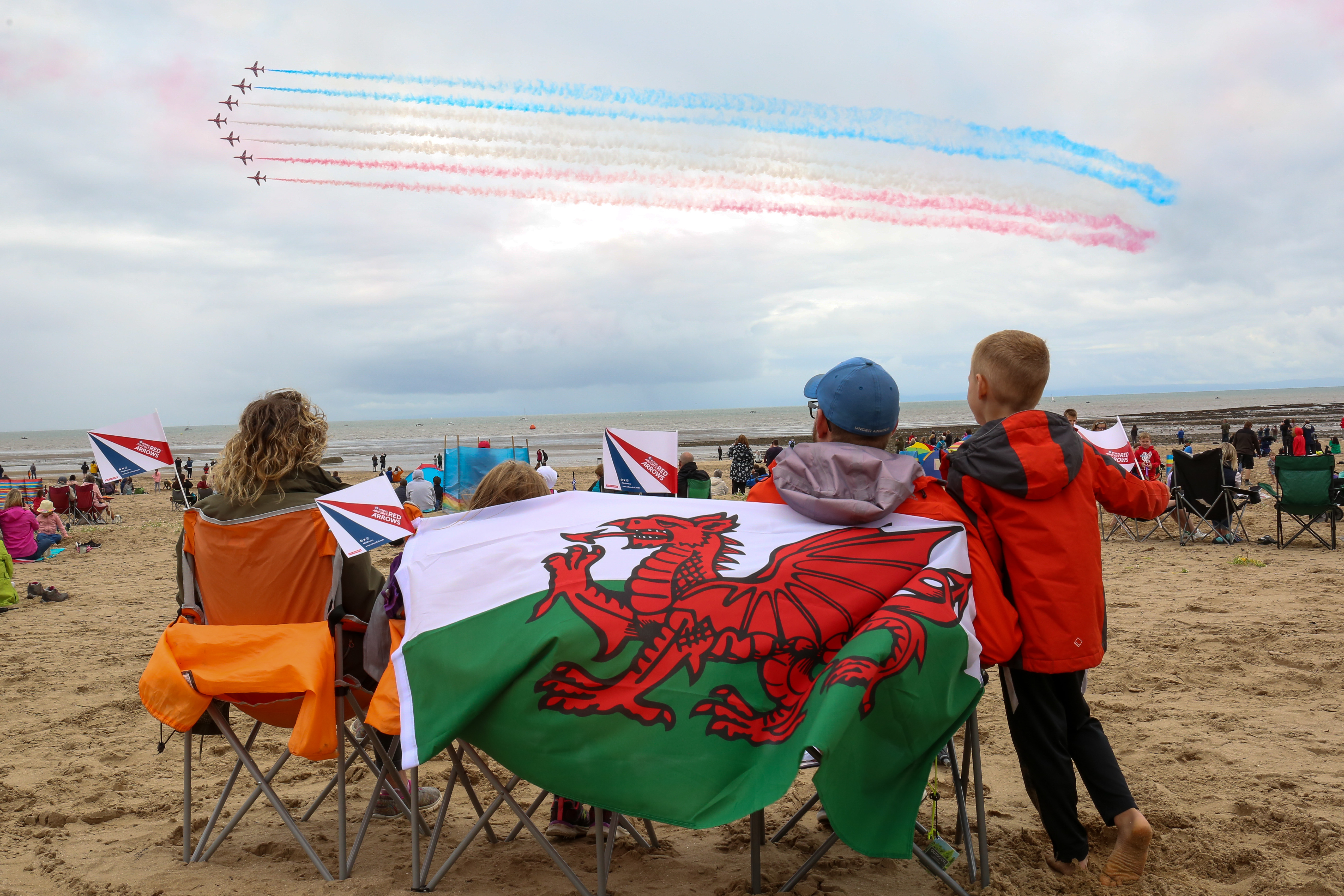Image shows the Red Arrows perform with red, white and blue colour trails, over a beach. Family watch from deck chairs, with a Welsh flag.