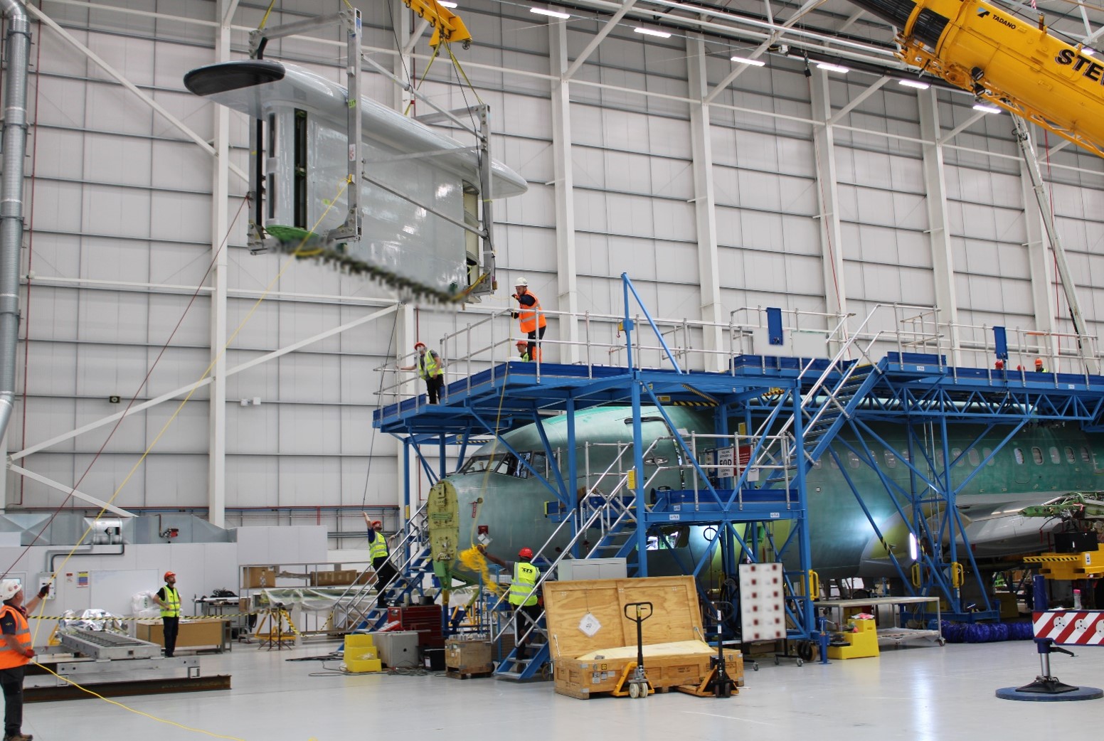 Image shows aircraft parts being assembled inside hangar. with crane lifting parts.