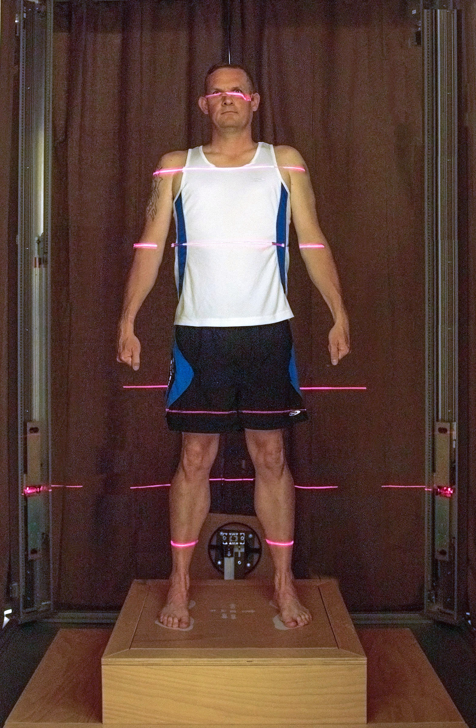 Image shows an aviator standing on a wooden block inside the body scanner, with scanning lines shining on his body.