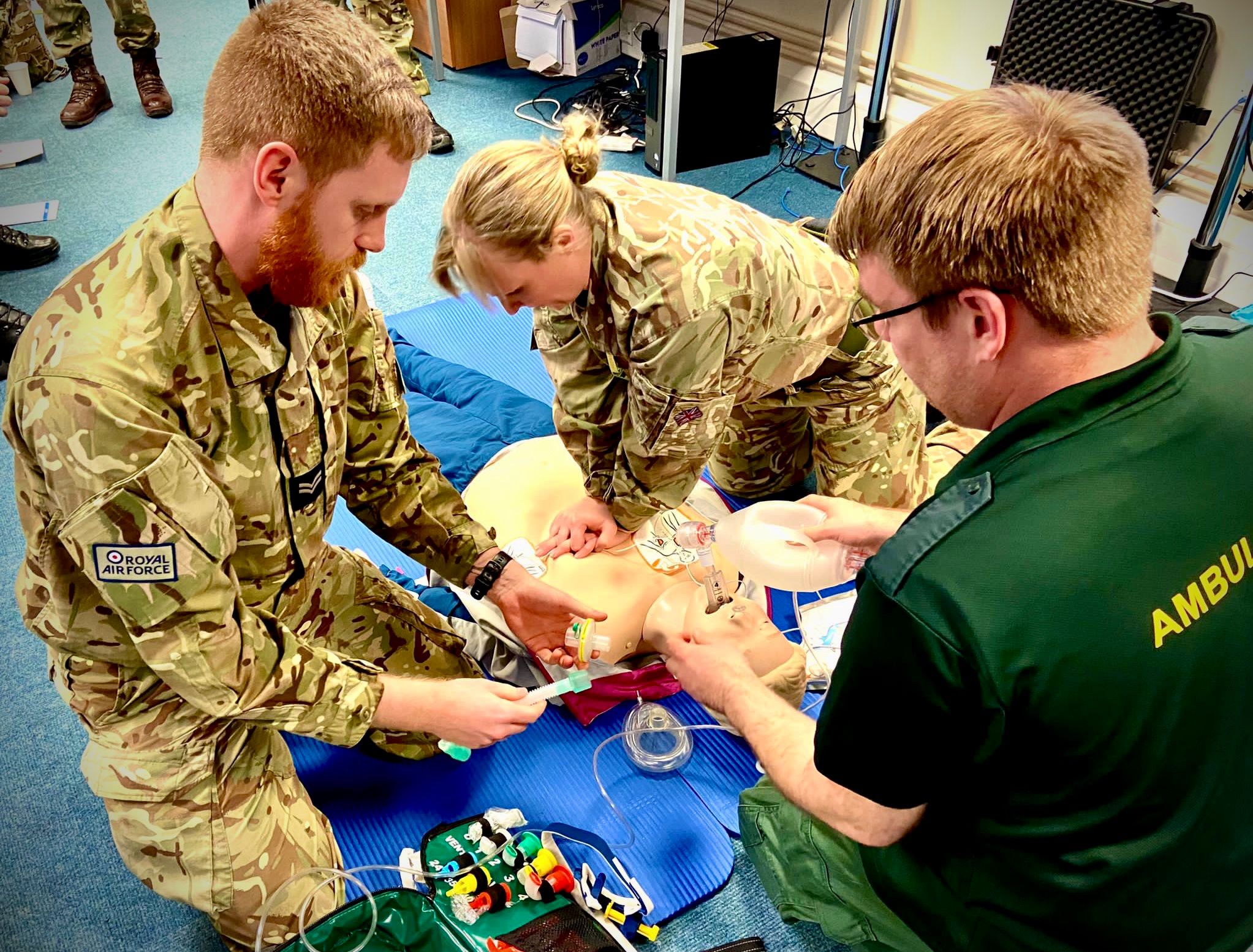 Image shows RAF Personnel and ambulance staff giving chest compressions to dummy in improvised classroom training scenario.