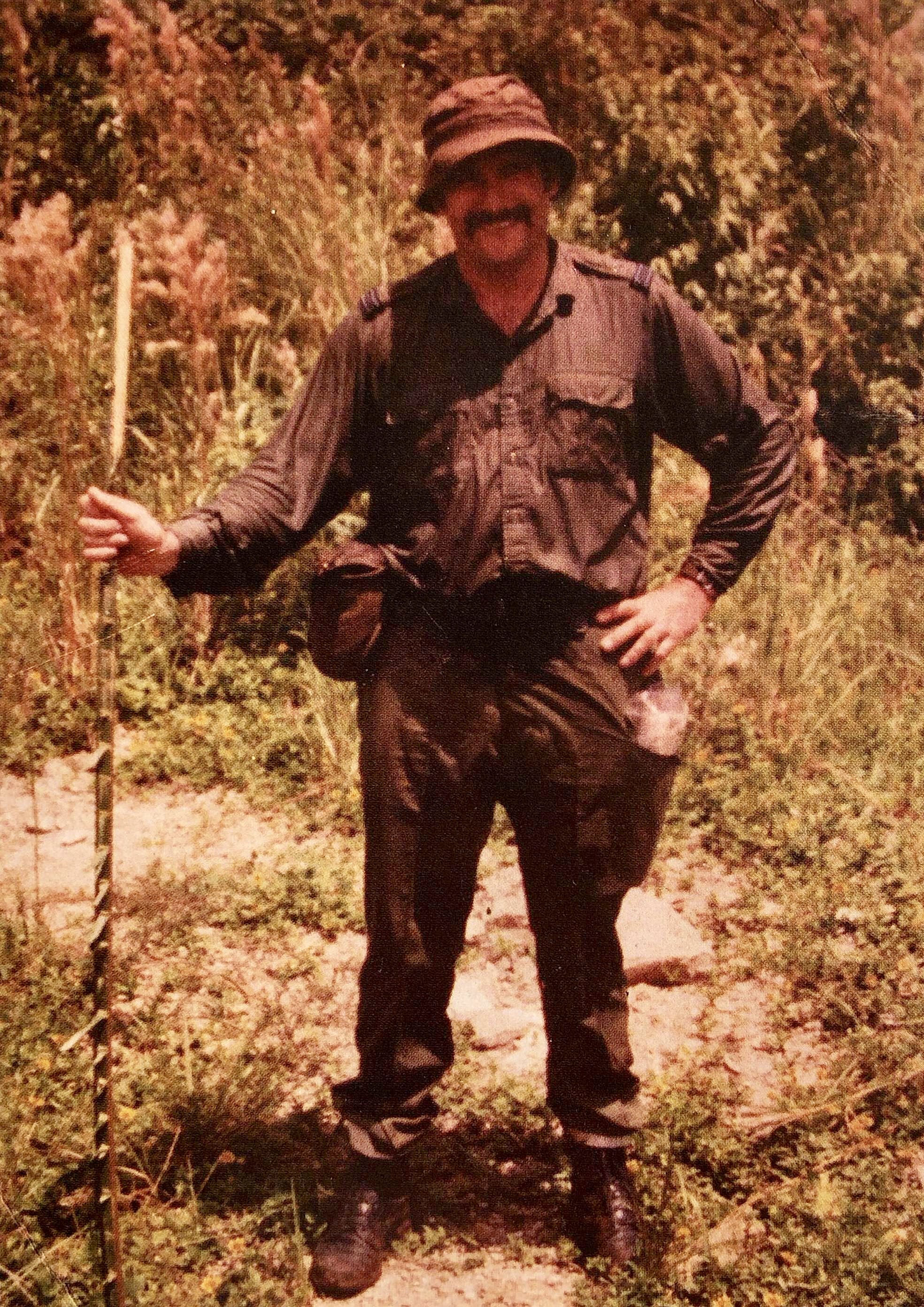 Old photo of soldier in grassy area holding walking stick.