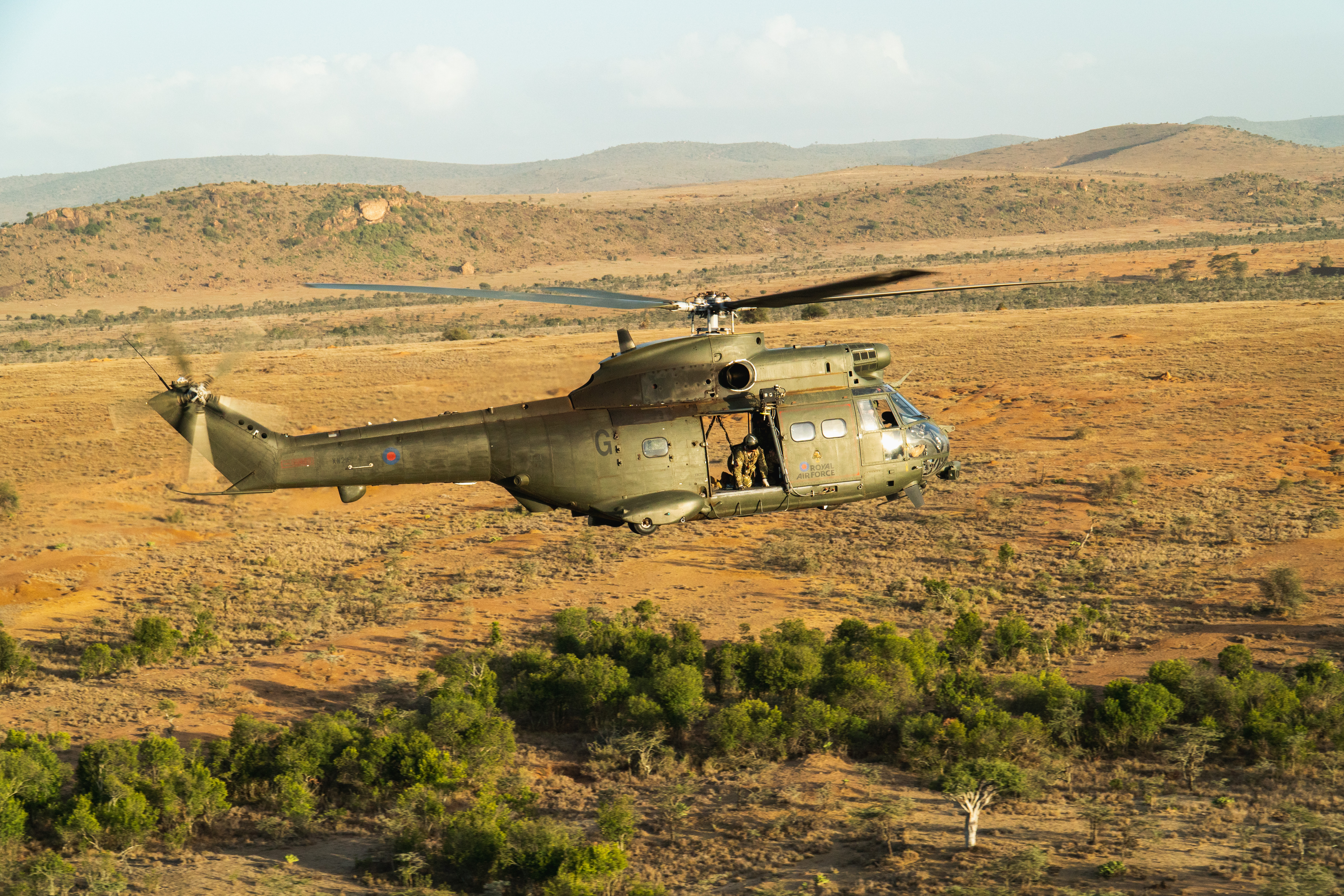 A Puma helicopter transits at low level across the desert, with the crewman visible in the side door looking at the small sections of greenery visible on the ground in amongst the desert.