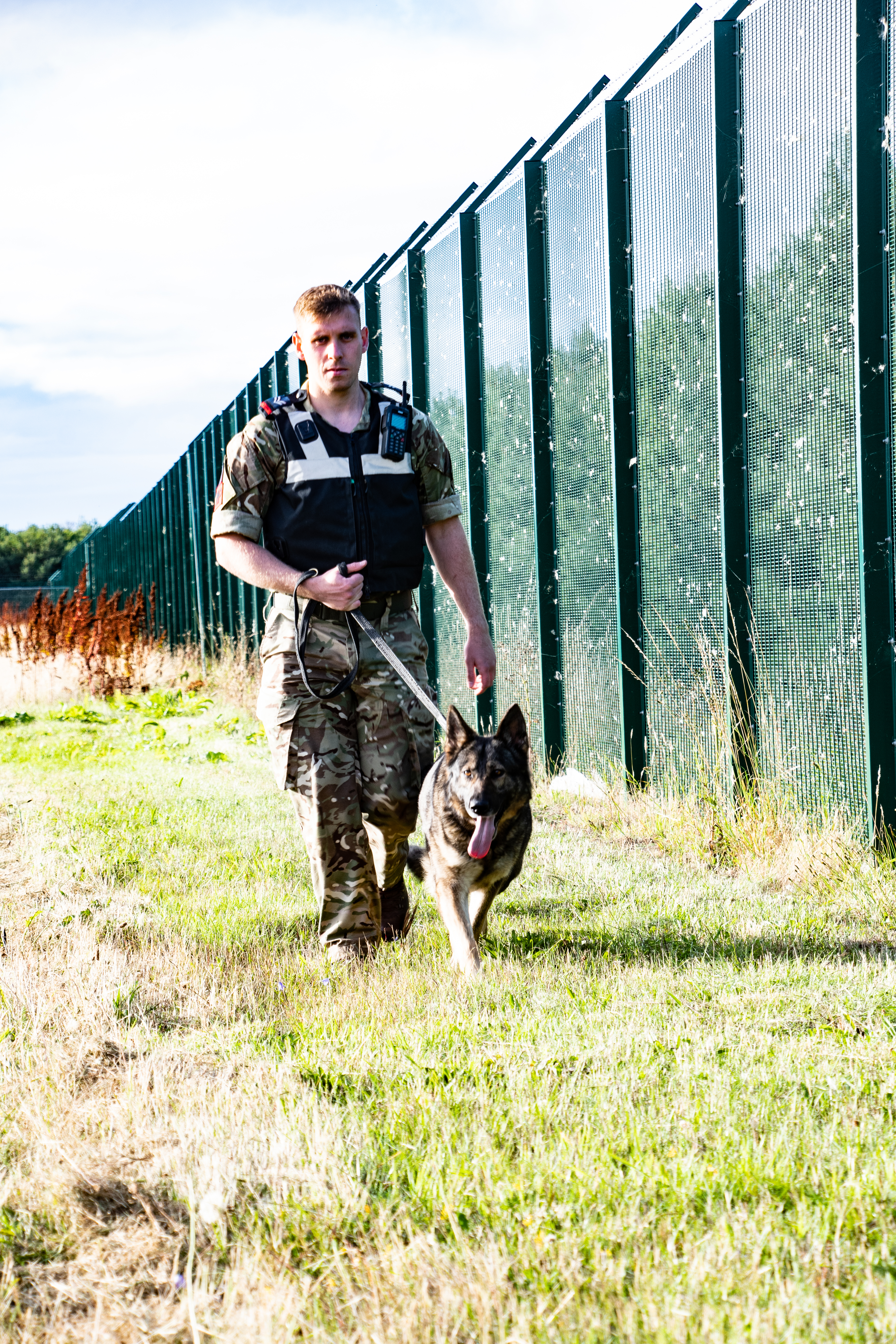 RAF Police Handler and dog walk by metal fence.