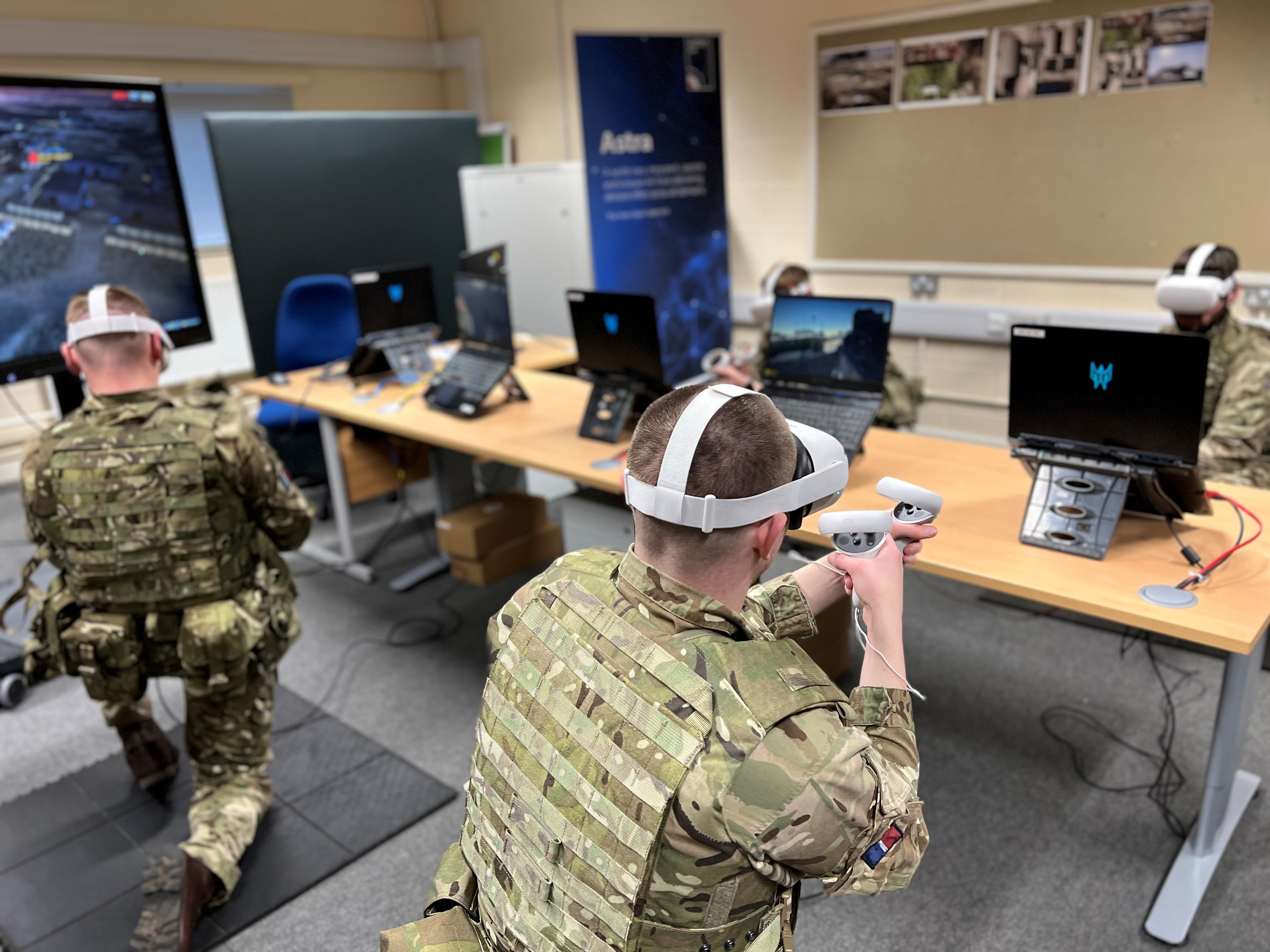 Personnel wear Virtual Reality headsets and use equipment. With computers and desk in background.