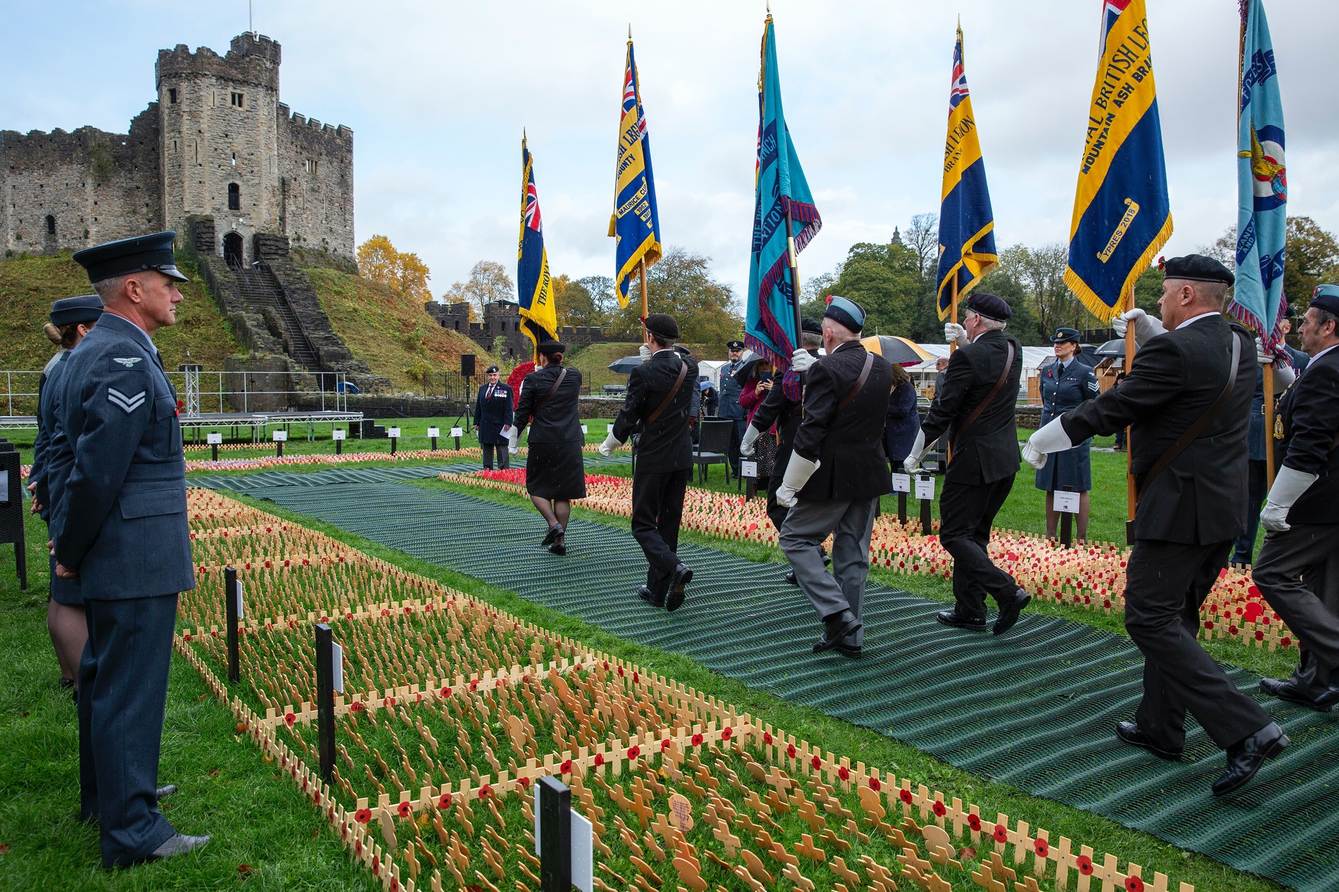 Image shows military personnel march with flags between rows of a poppy crosses towards castle.
