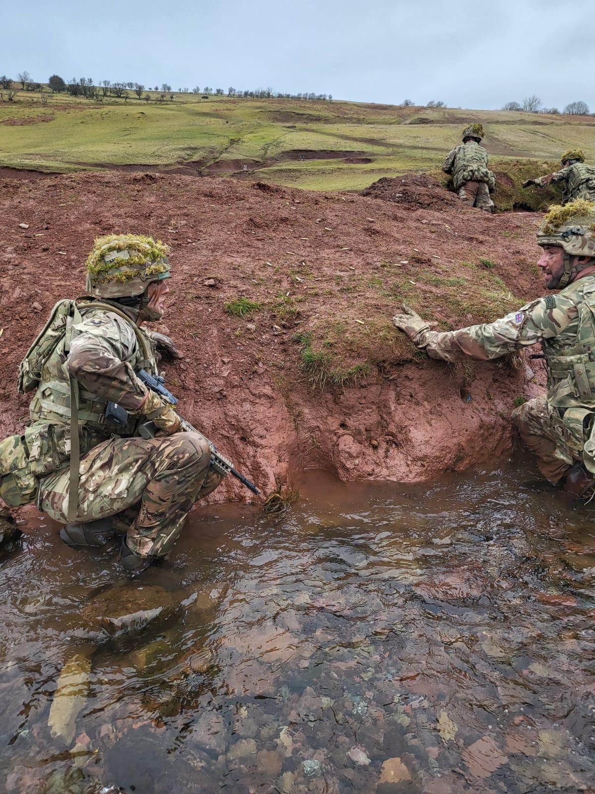 Personnel crouch in river by muddy bank.