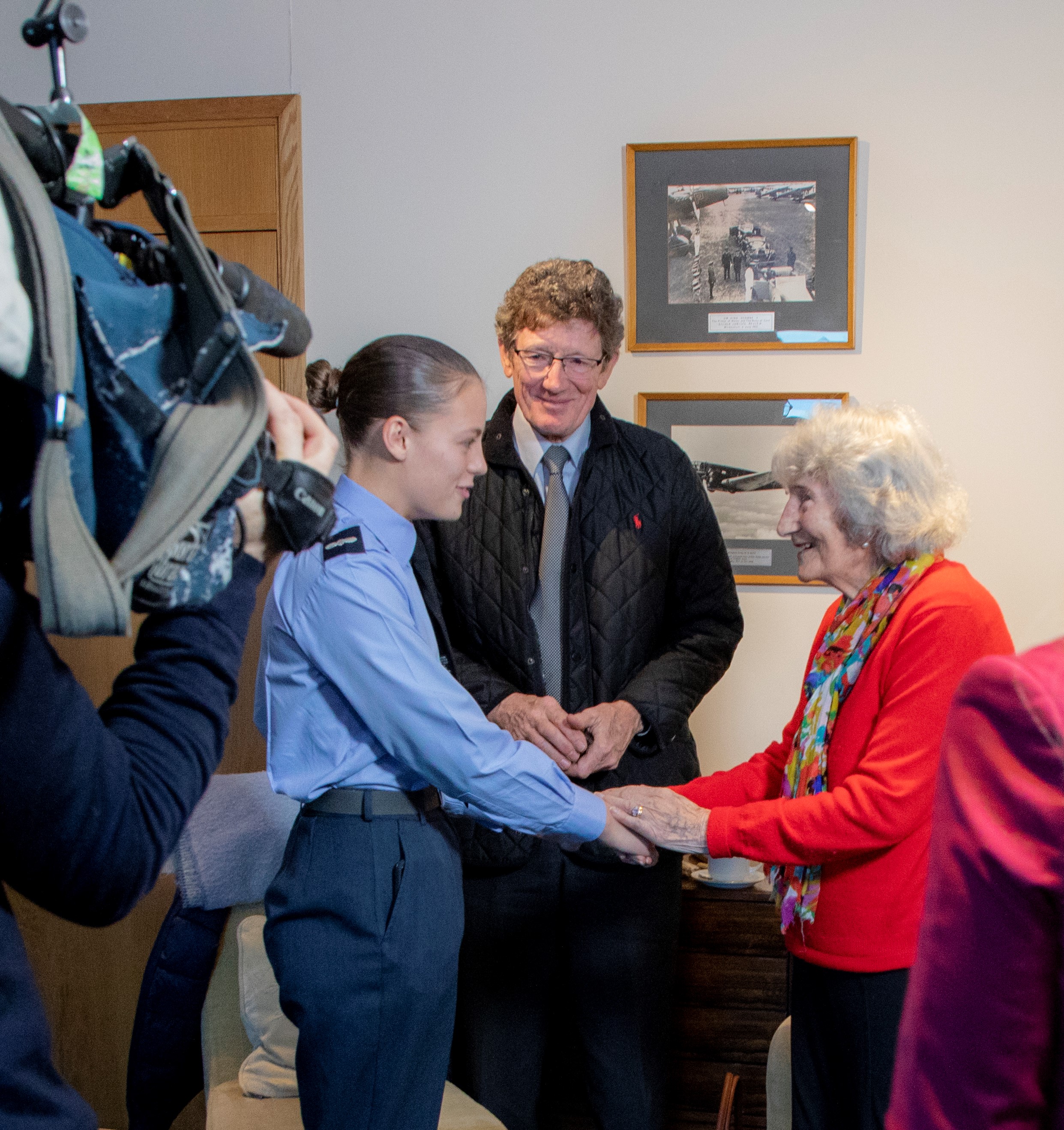Image shows Veteran shaking hands with RAF aviators, as camera takes pictures.