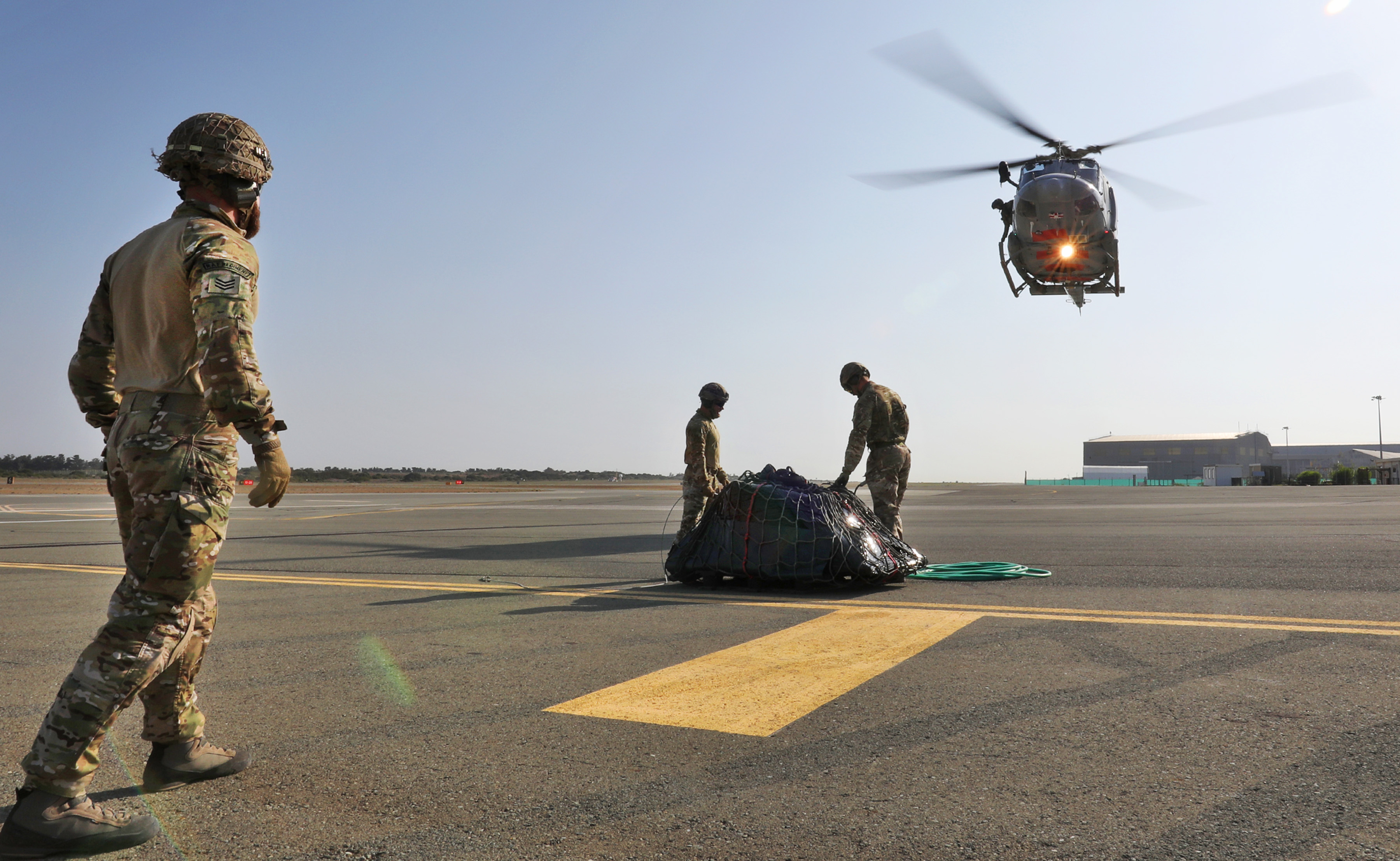 Image shows RAF aviators with cargo on an airfield, as a Chinook helicopter flies overhead.