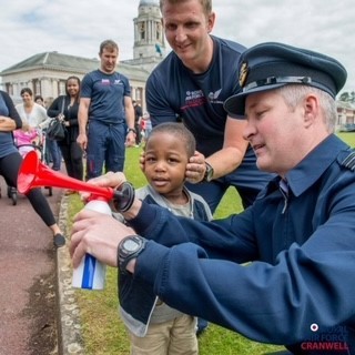 Image shows RAF aviator sounding a klaxon as another covers the ears of a child.. 