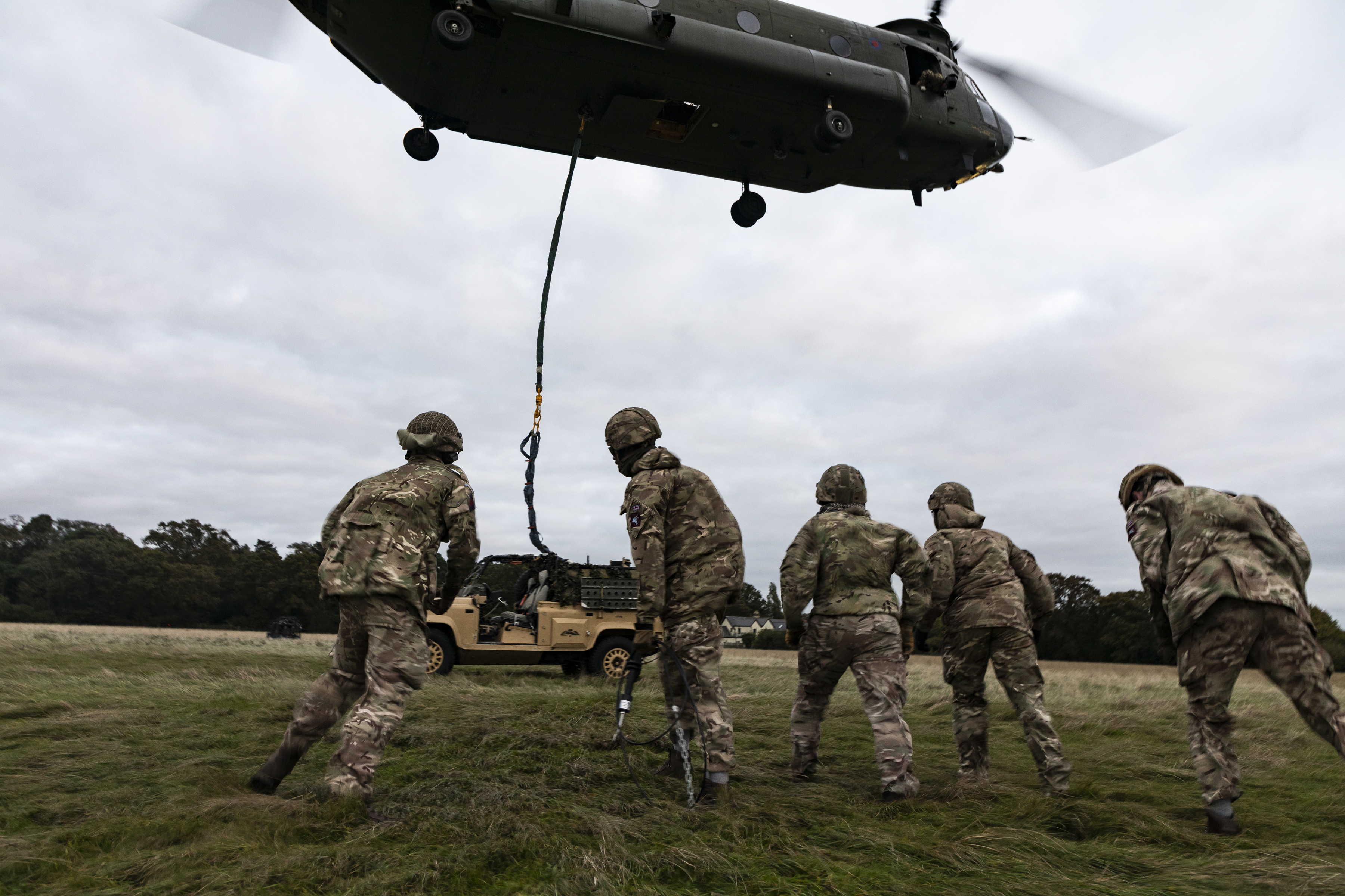 Soldiers from 23 Parachute Engineer Regiment standing under a hovering Chinook while they hook a vehicle underneath for carriage