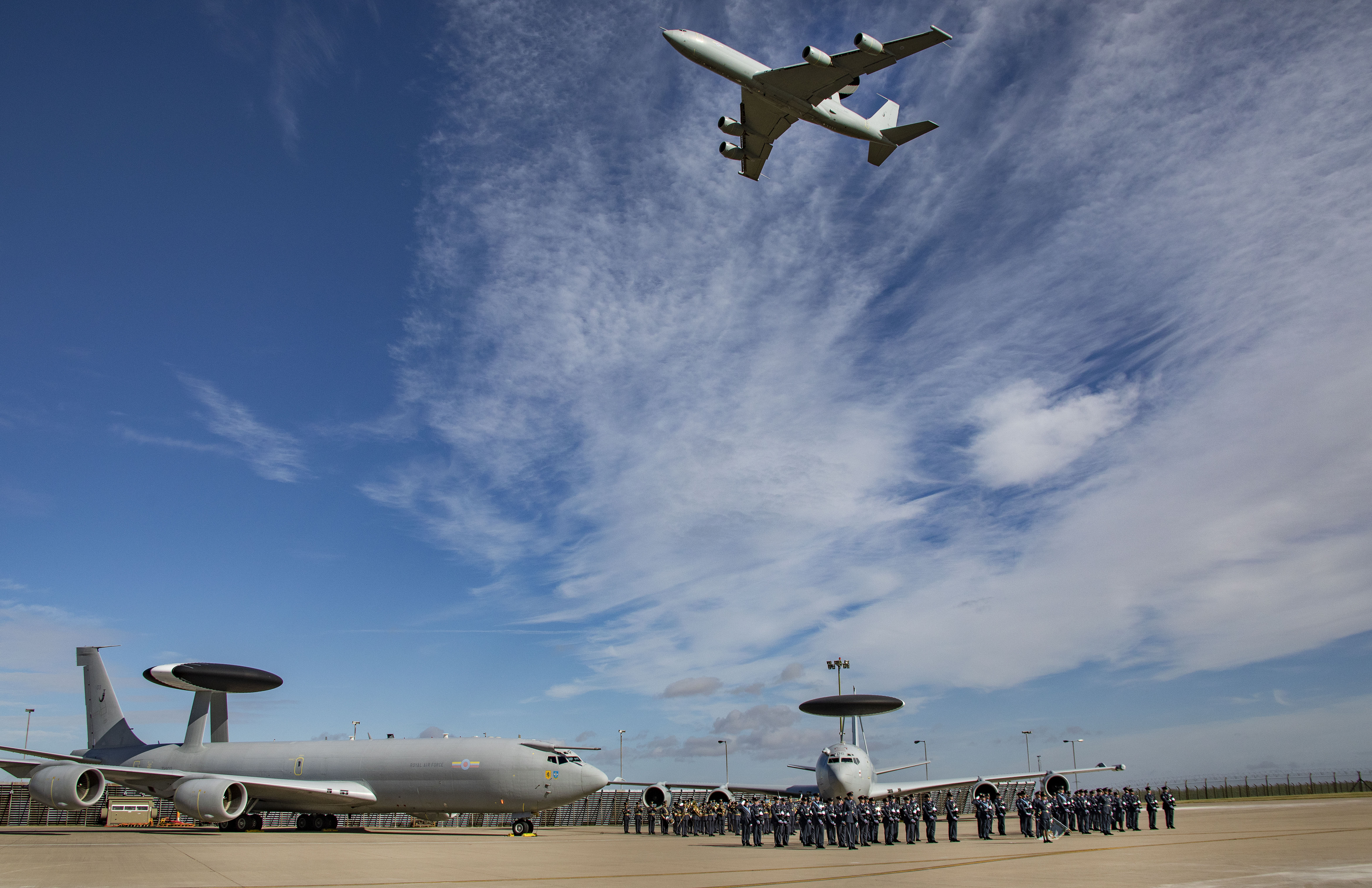 Two aircraft on the runway with parade, as aircraft flies over.