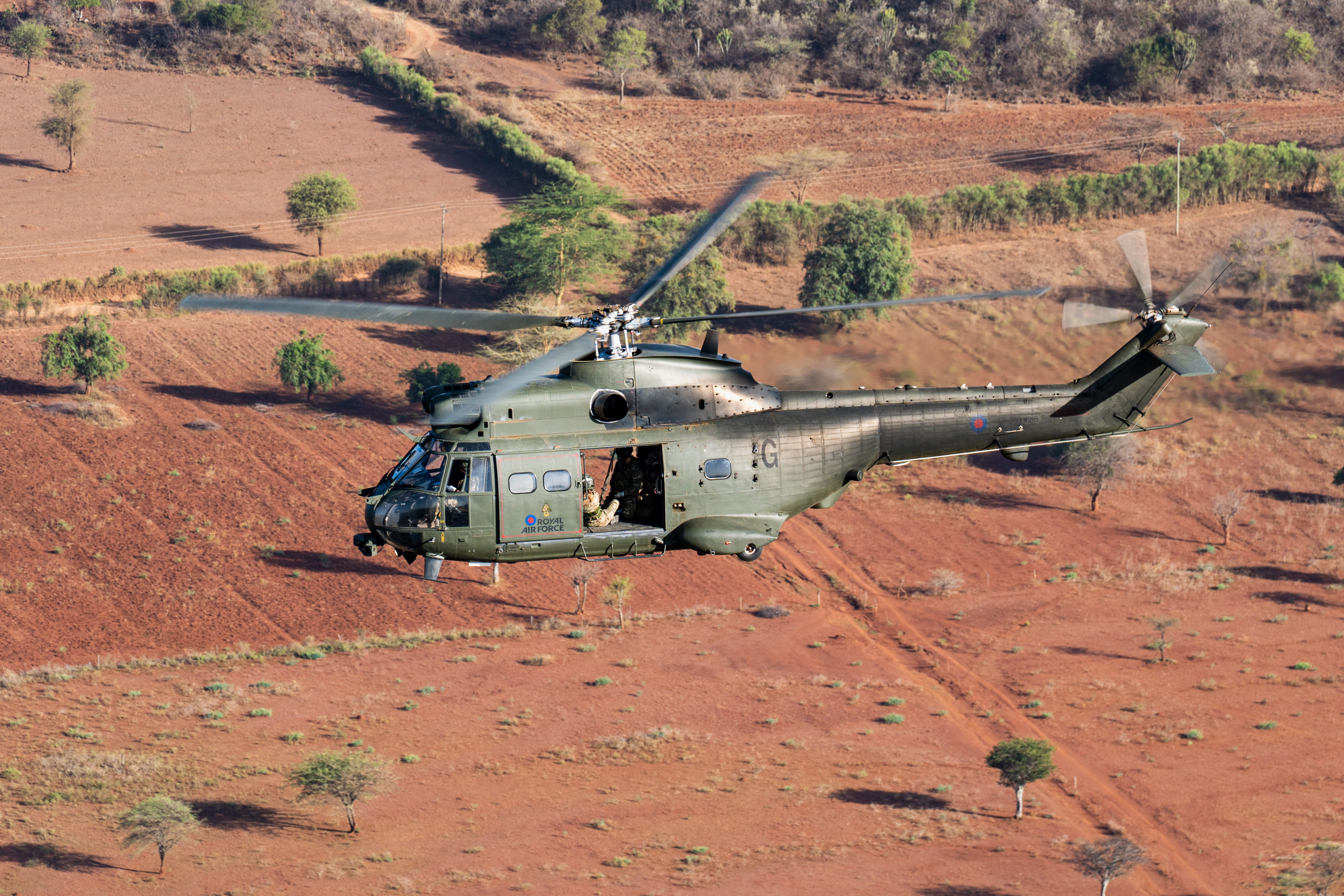A Puma helicopter flies at low level across the Kenya desert, with small lined areas of trees visible amongst the almost red-looking sand.
