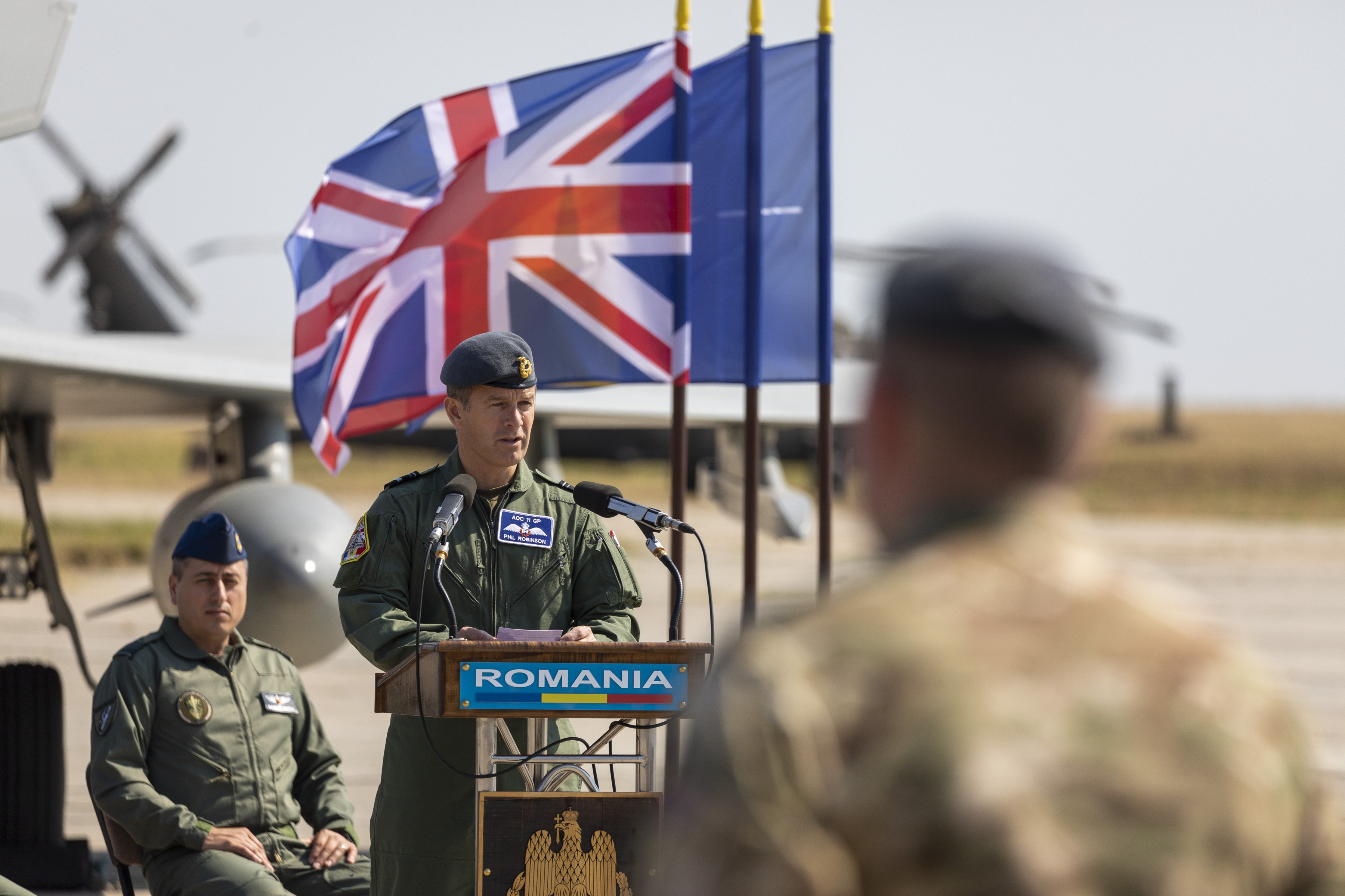 Image shows aviators giving presentation with aircraft and flags in the background.
