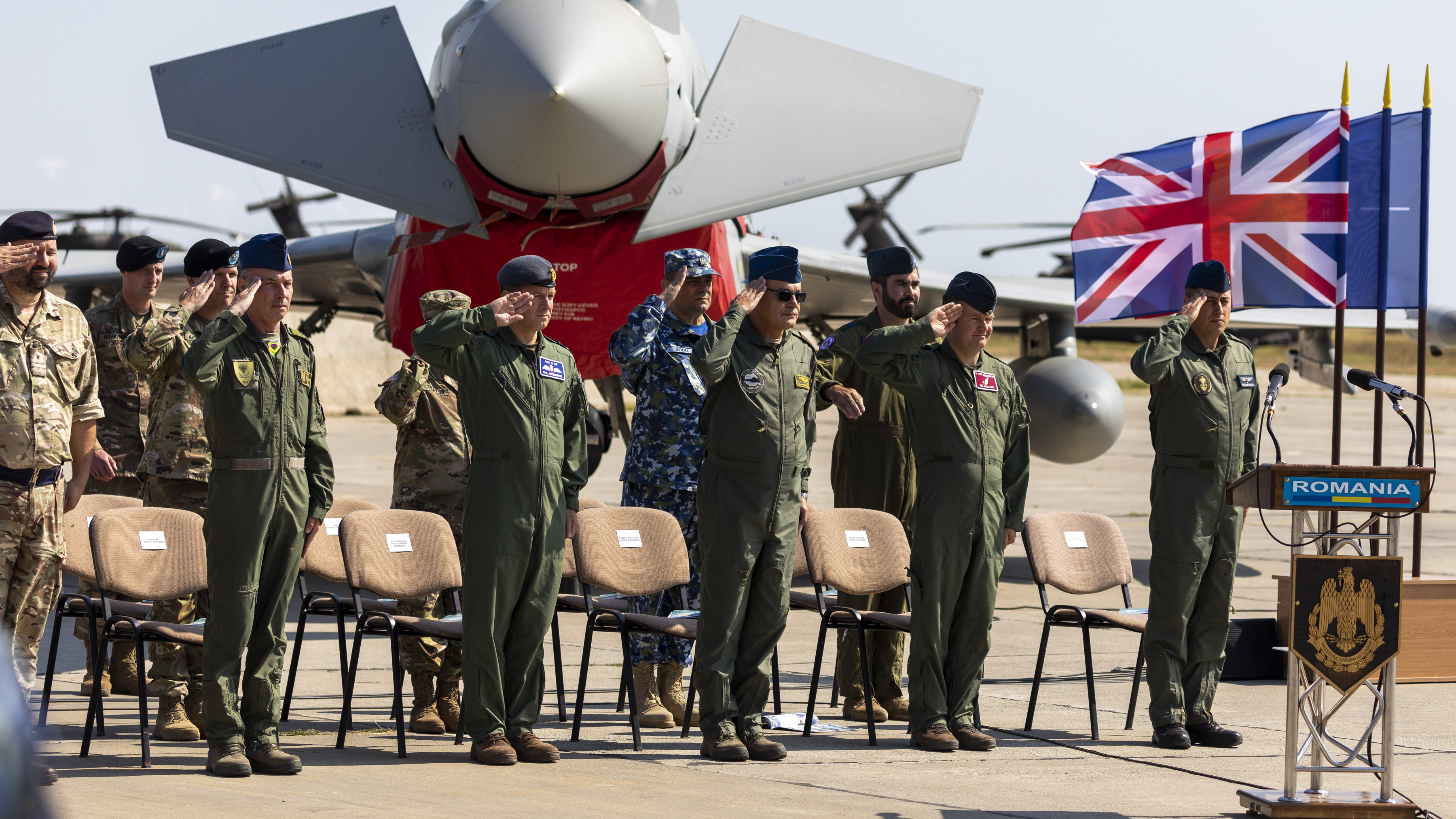 Image shows Squadron saluting by chairs with aircraft and flags in the background.