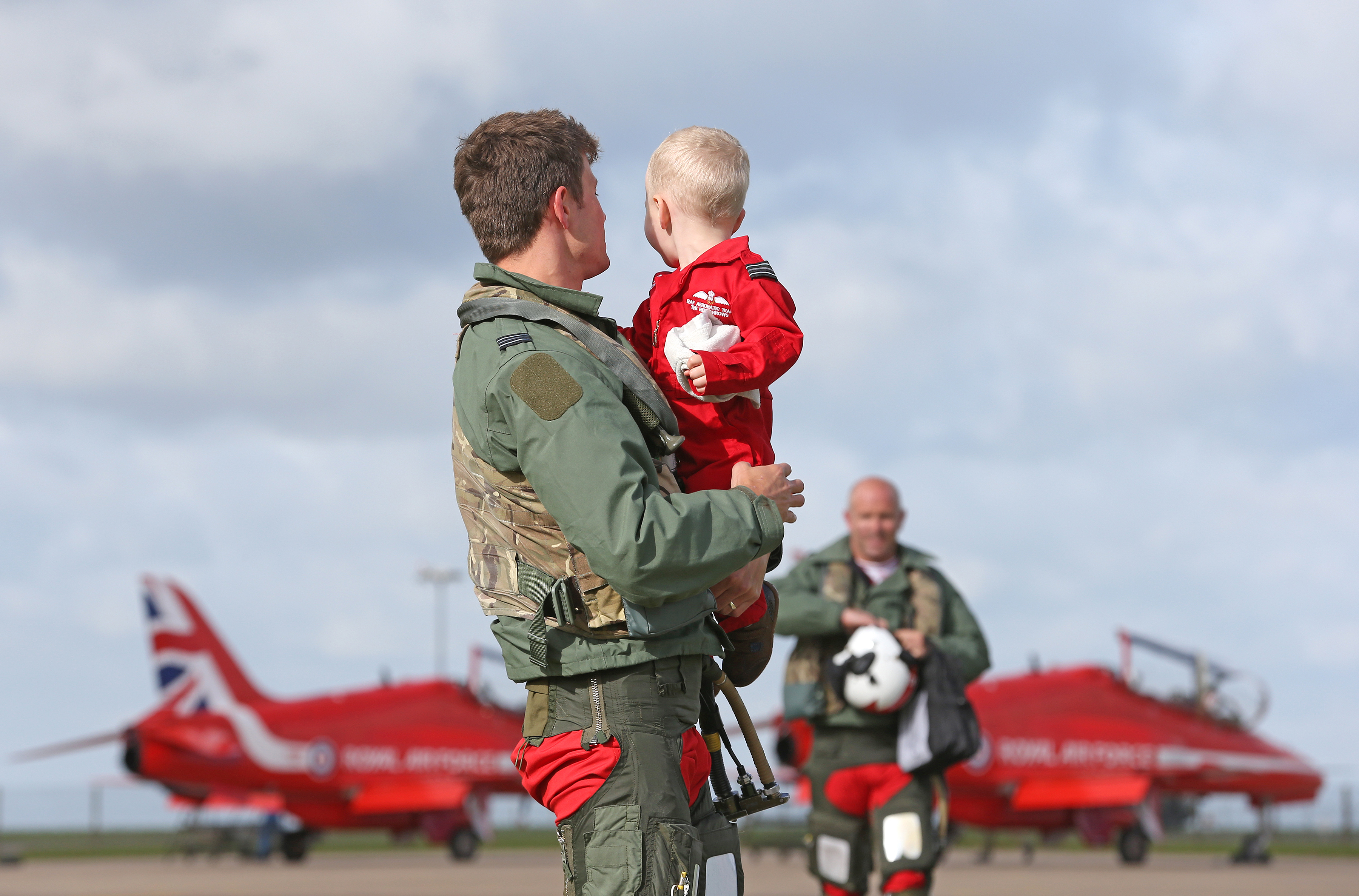 Red Arrow's pilot holds son, with another pilot and two Hawk T1's in the background.