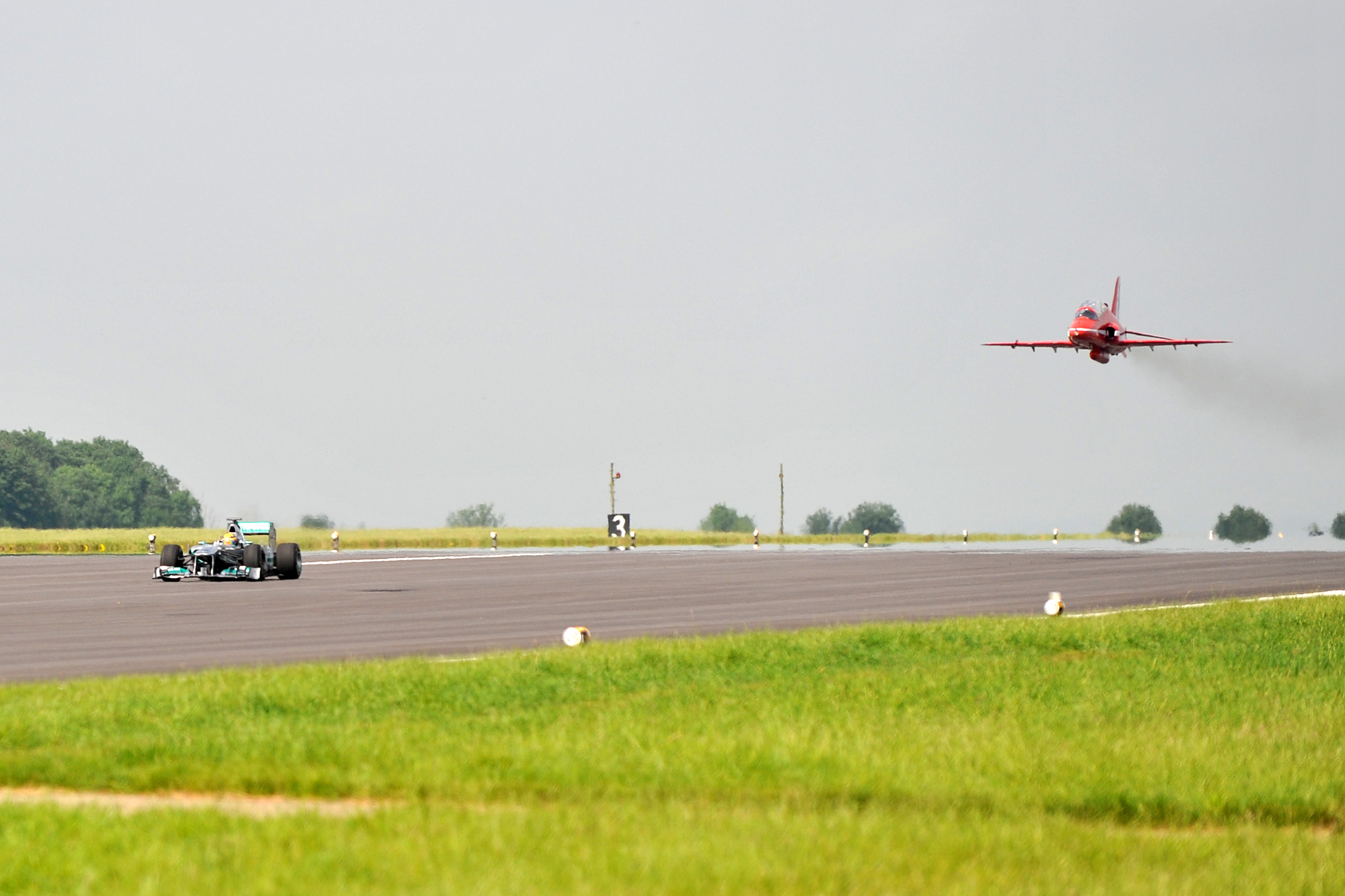 Lewis Hamilton drives his F1 car along the runway, with a Red Arrows jet overhead at RAF Scampton in 2013.