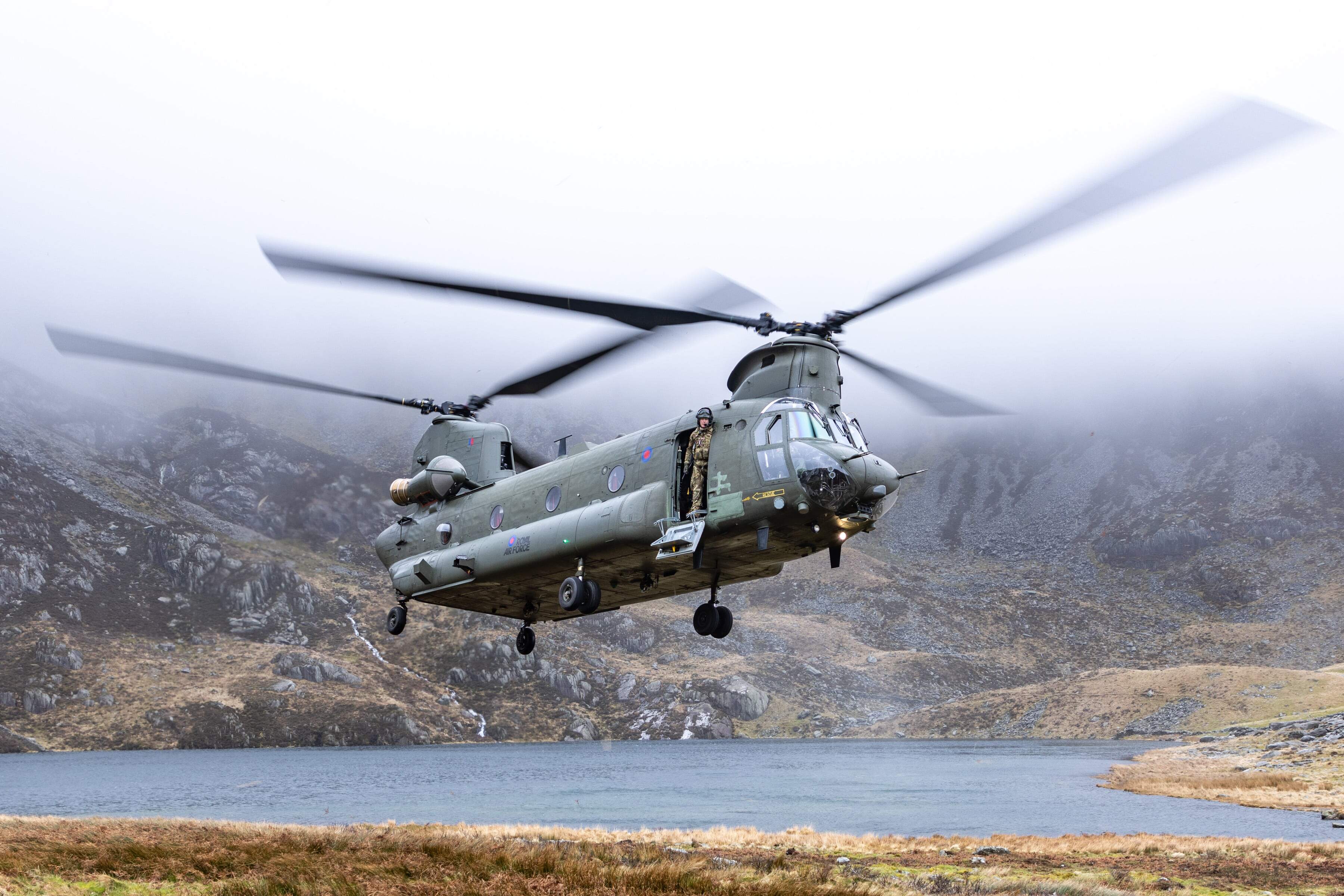 Chinook hovering low over a mountain lake with someone standing on the edge of the doorway