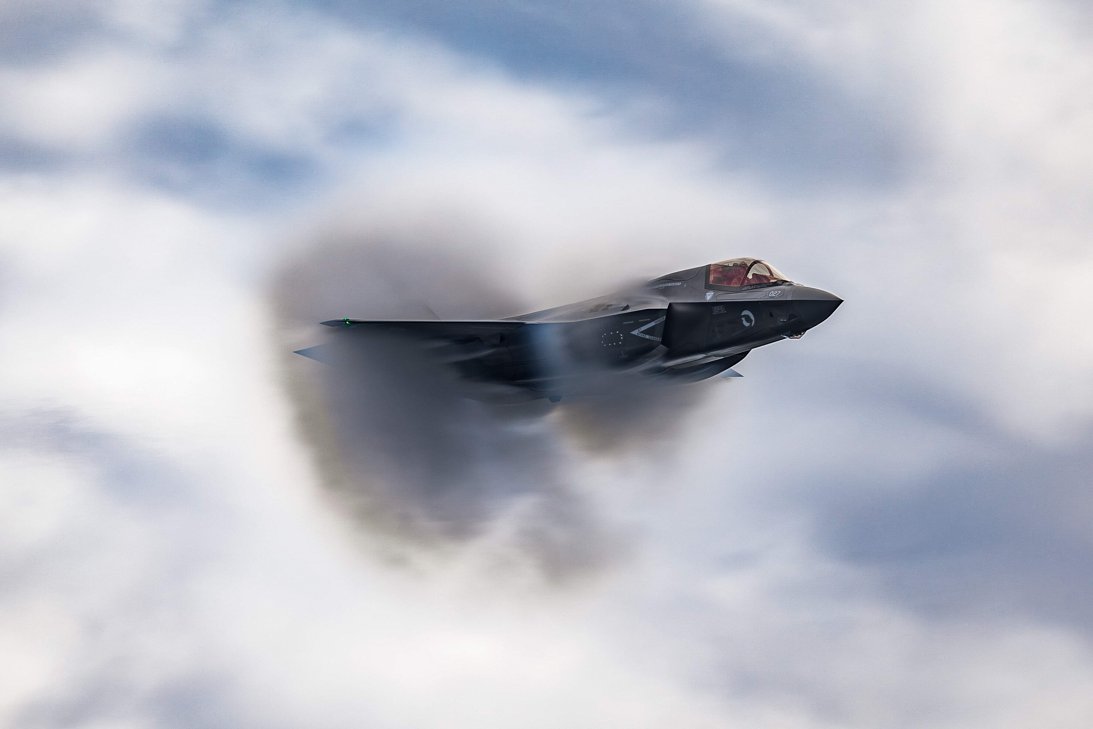 F-35B aircraft flying against a cloudy sky creating a vapor cloud behind it