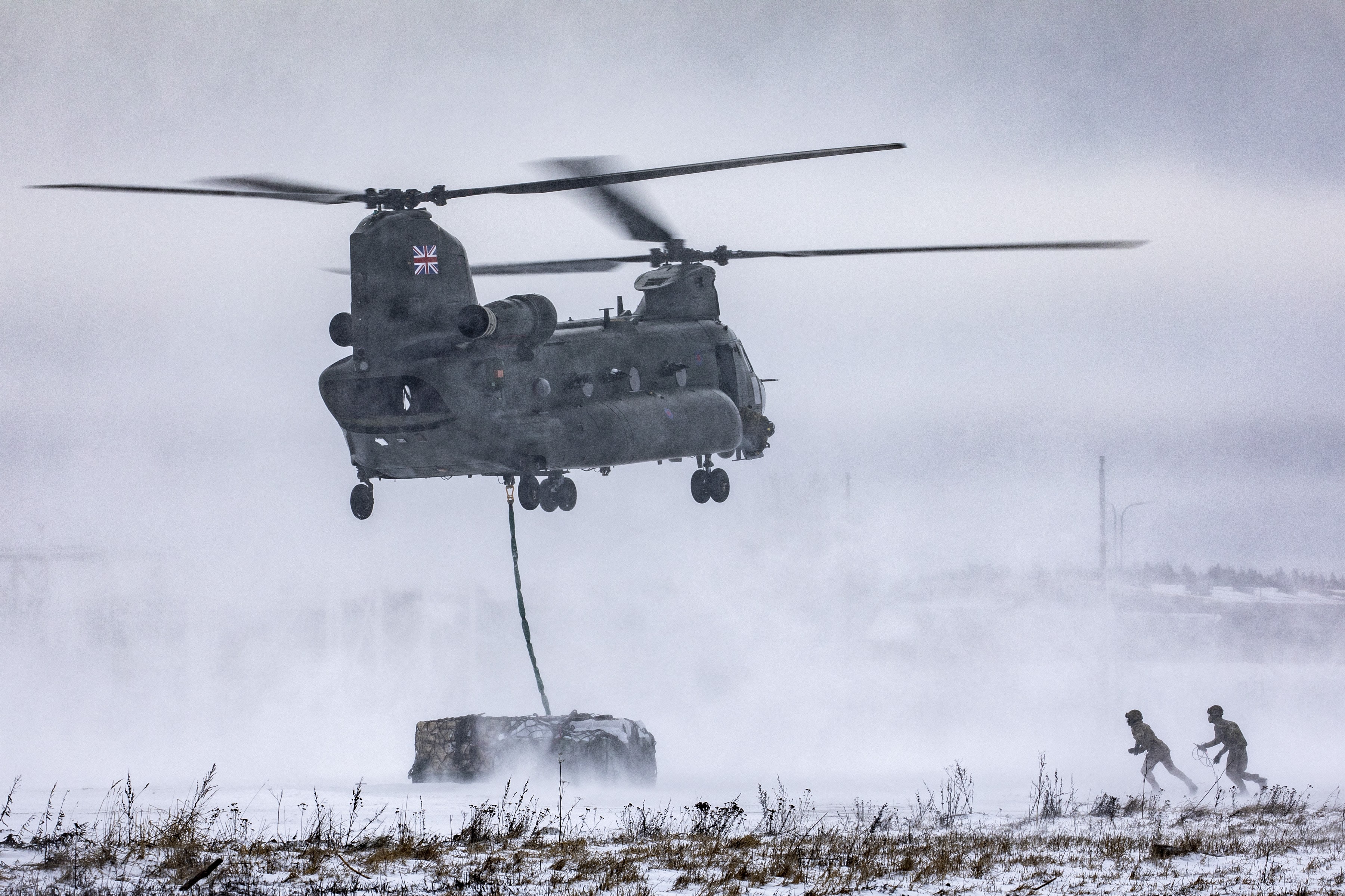Chinook hovering over snowy ground, landing an underslung load, with 2 RAF personnel running to unclip it. 