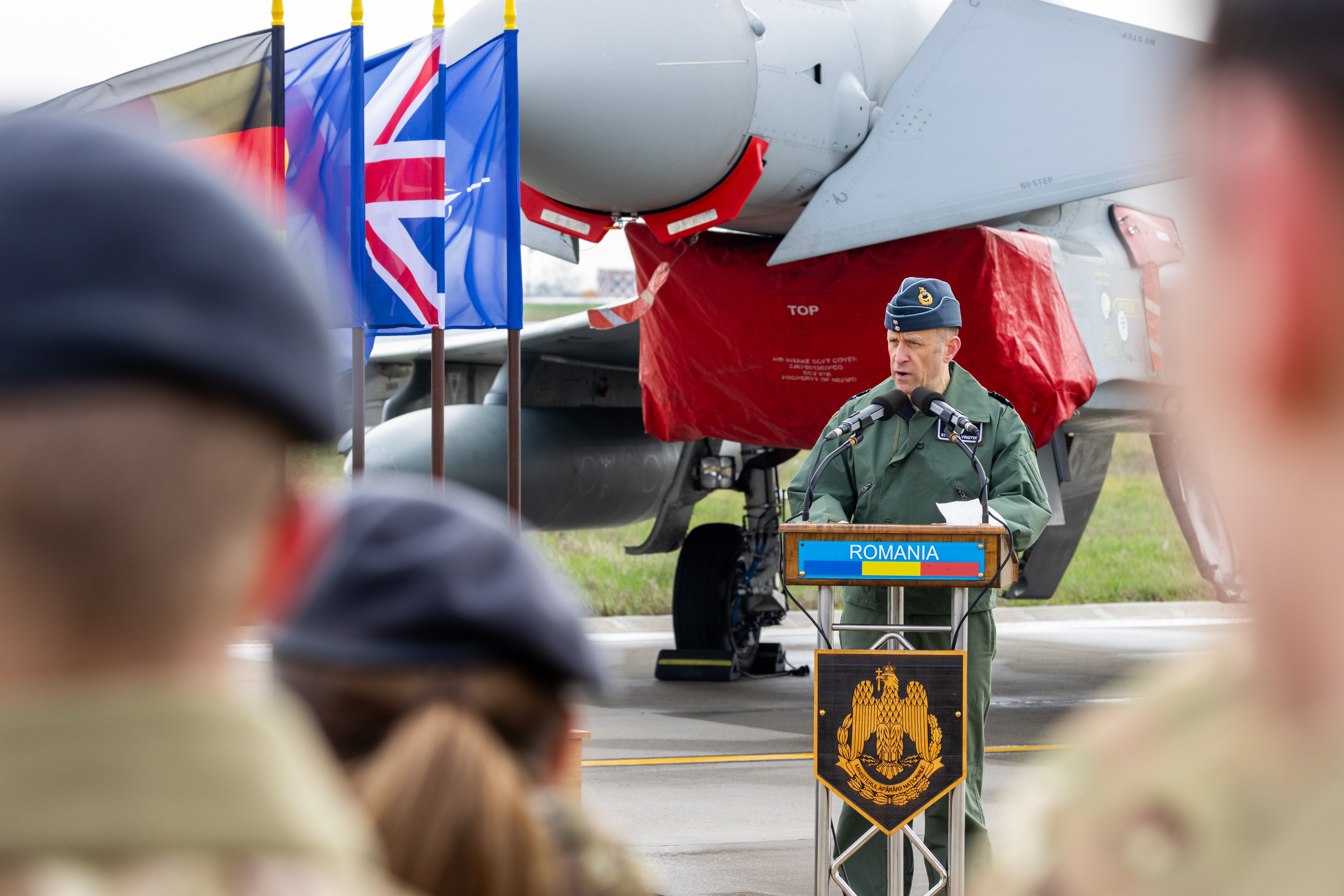 An Air Force Officer speaking at the handover ceremony in front of a Typhoon and allied flags.