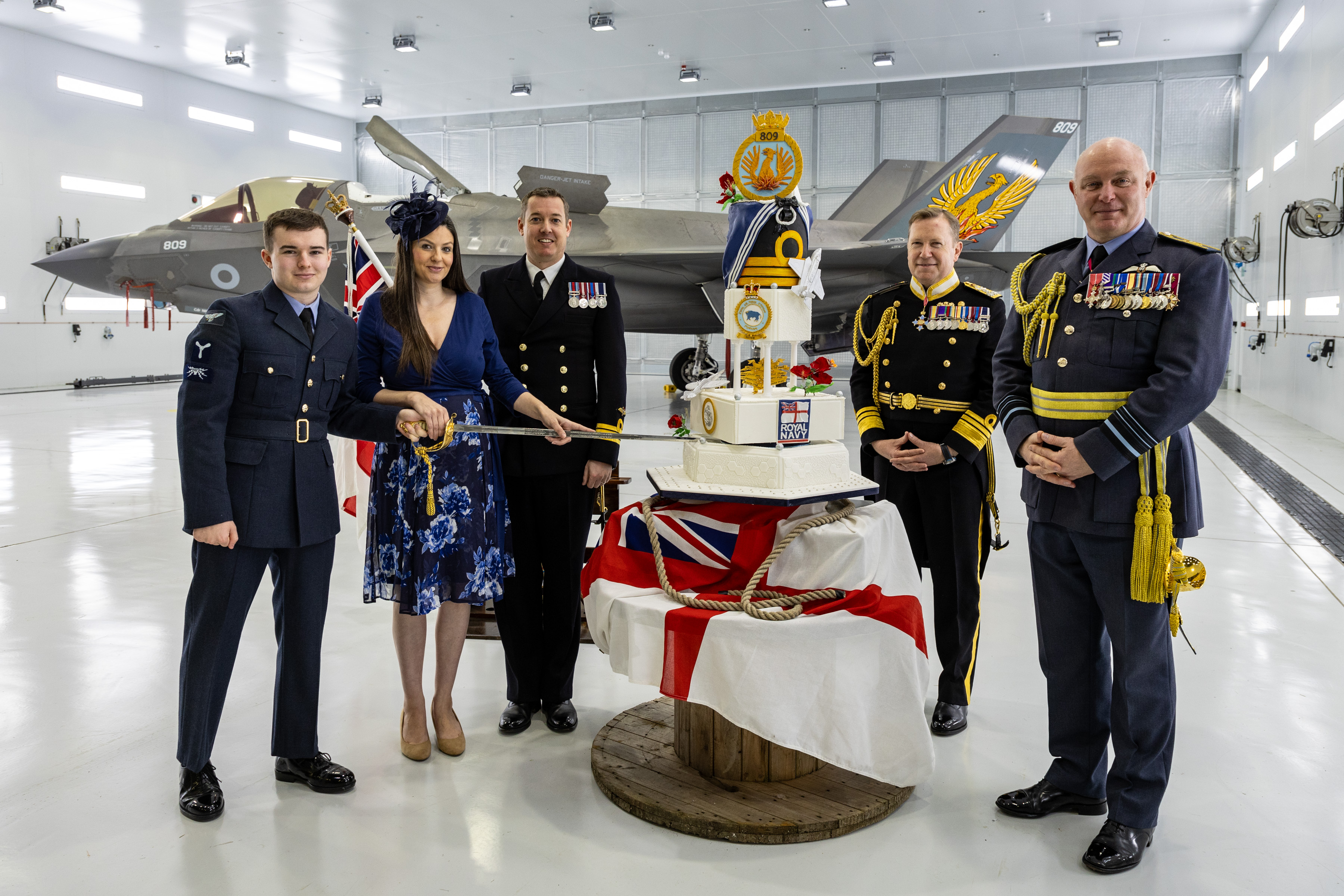 Group of 5 people from a mix of the RAF and the Navy standing facing the camera, with a 809 Squadron cake being cut in the middle.