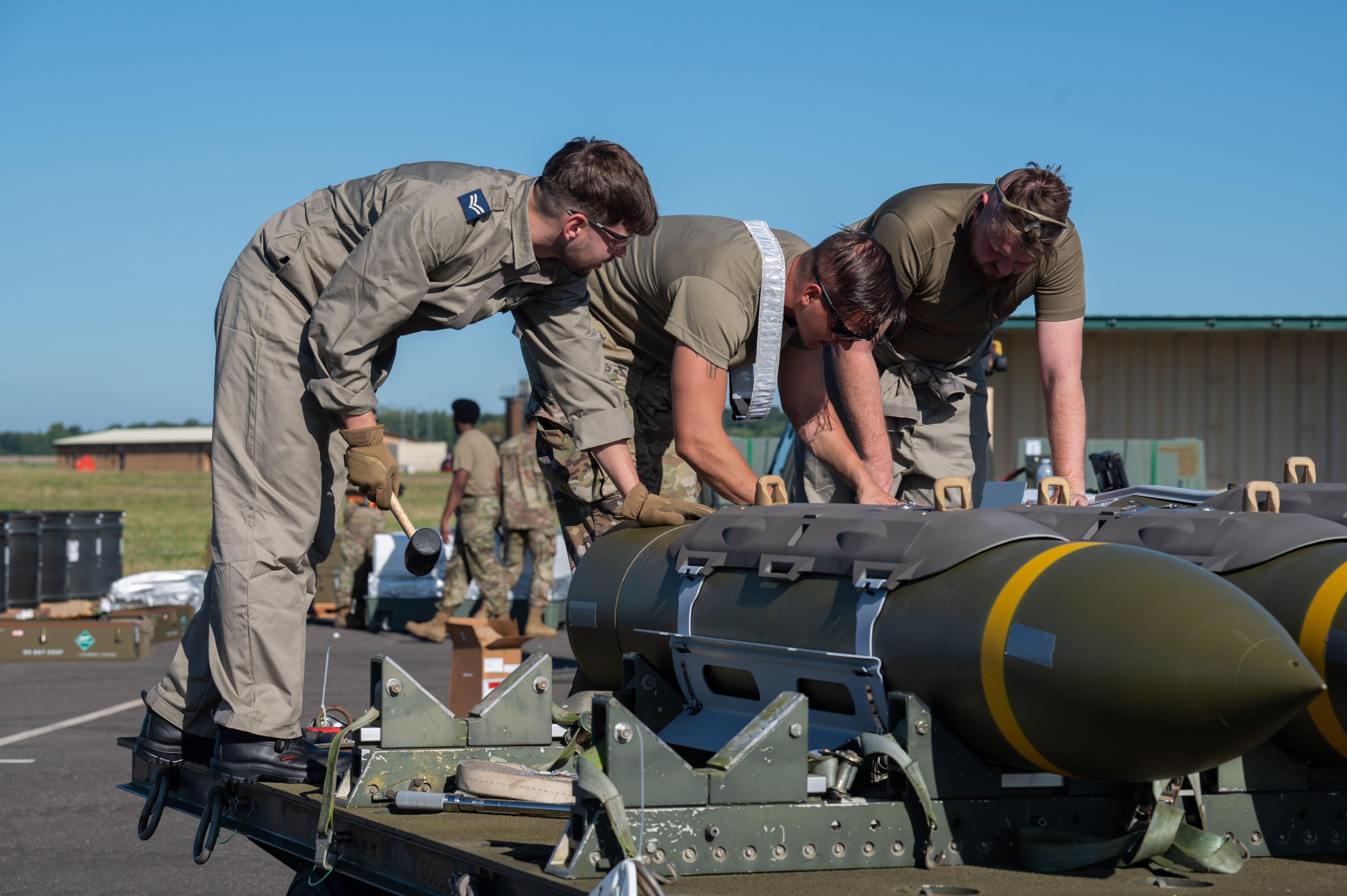 Military personnel working on armaments strapped to a trailer on an airfield