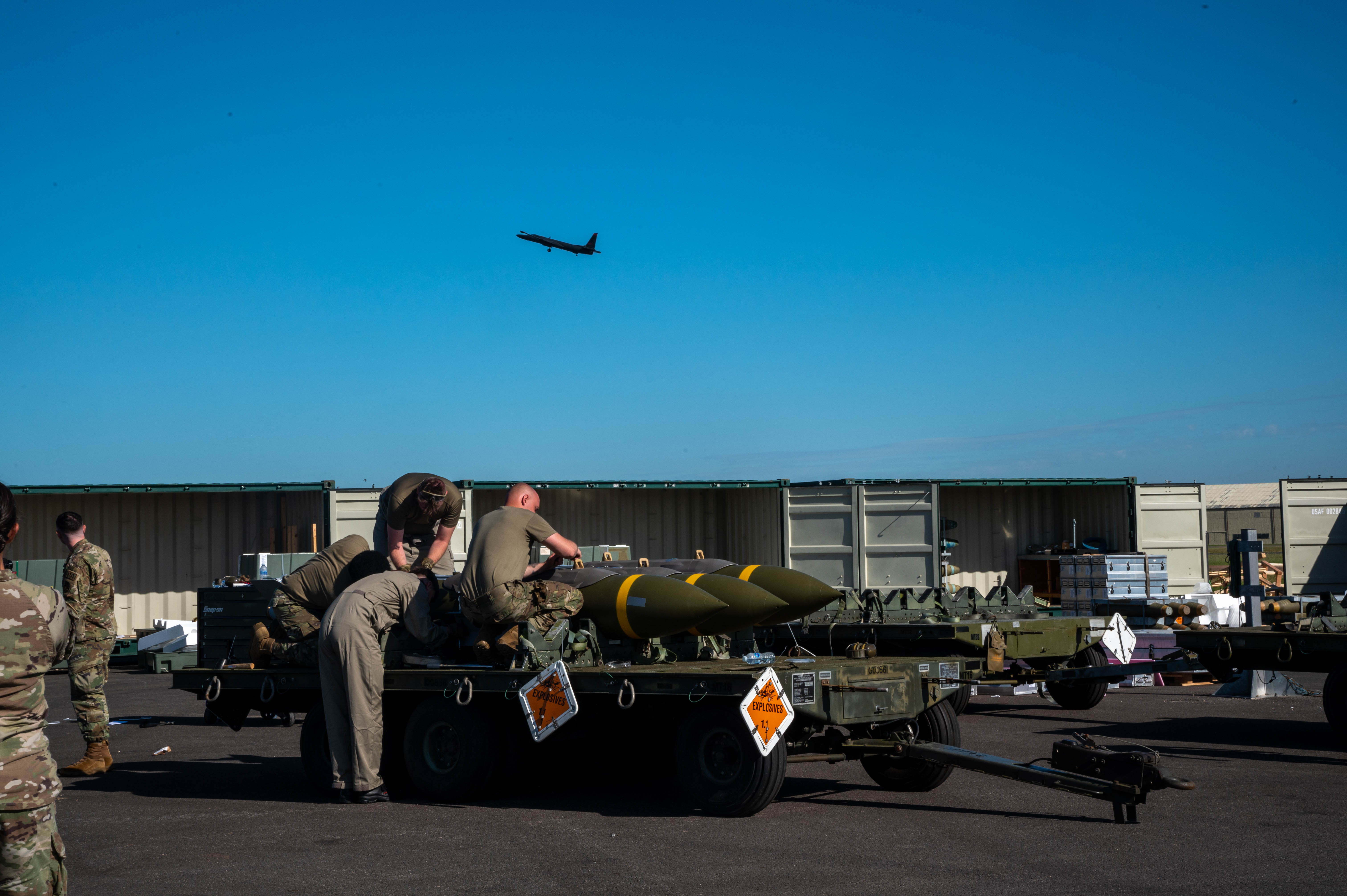 Military personnel strapping armaments to a trailer, with an aircraft flying overhead