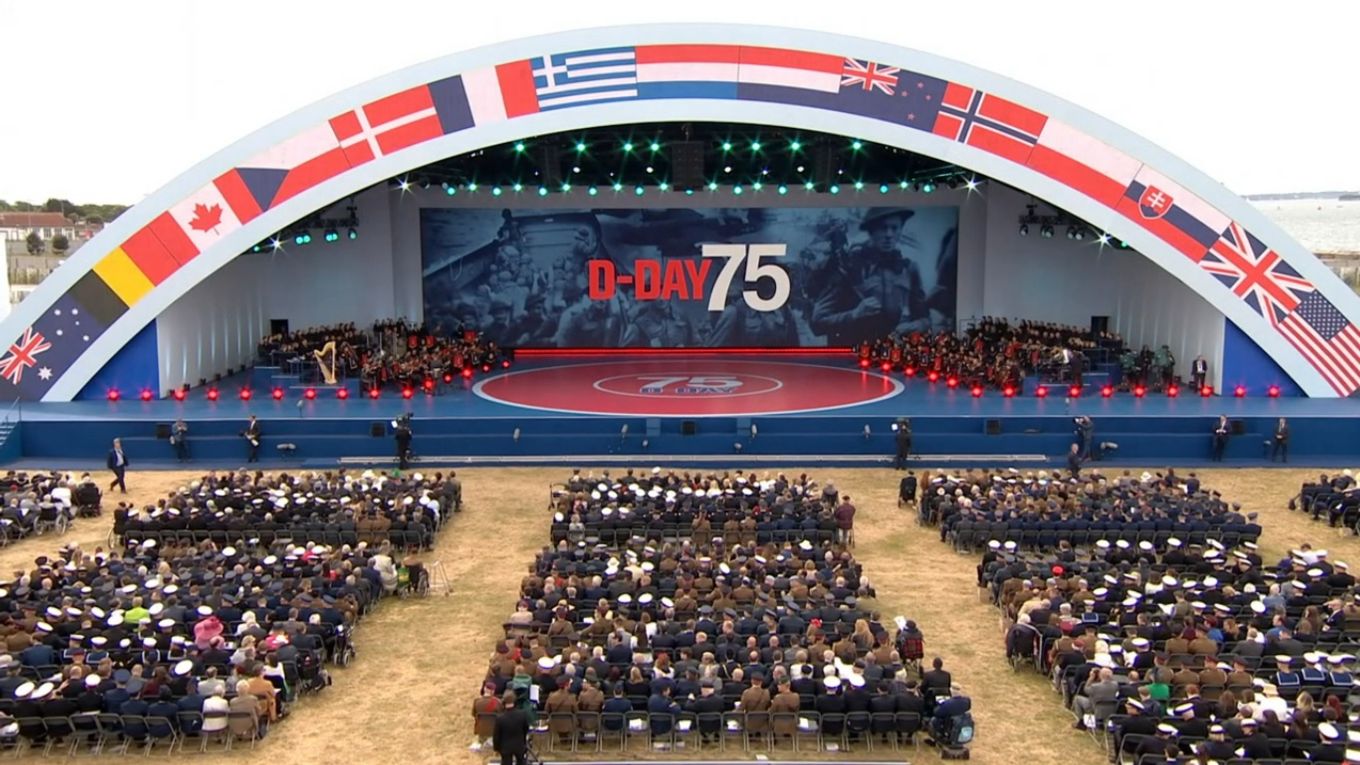 An audience of veterans watch the D-Day75 Commemorations take place on a large stage.