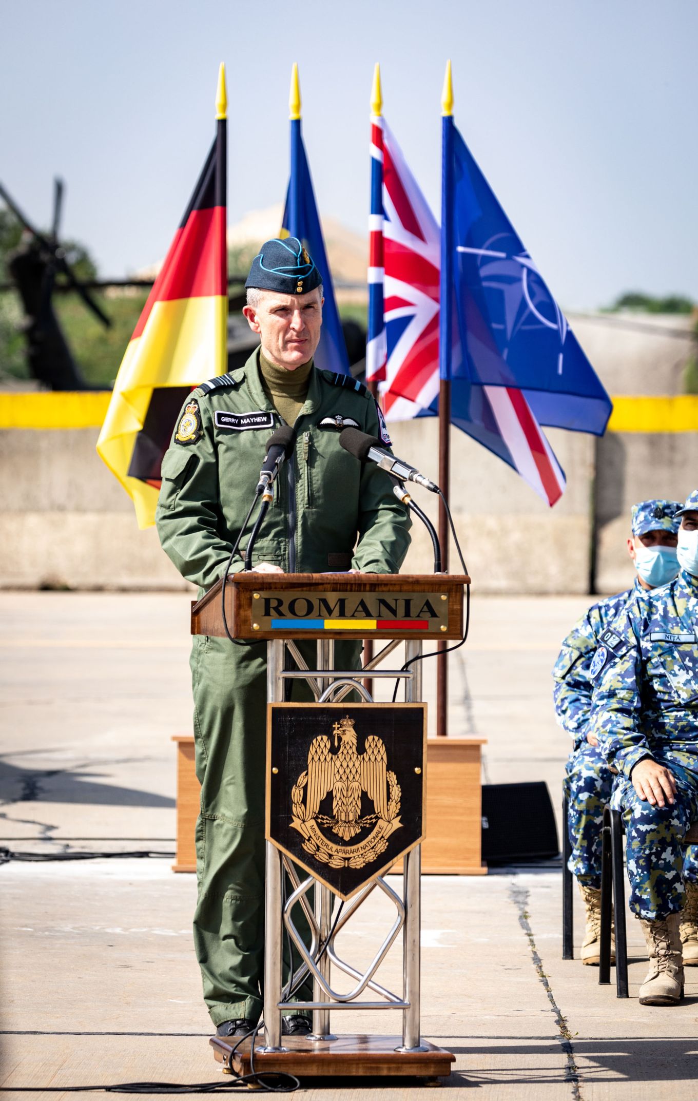 Air Marshal Mayhew standing at a microphone podium with flags behind.