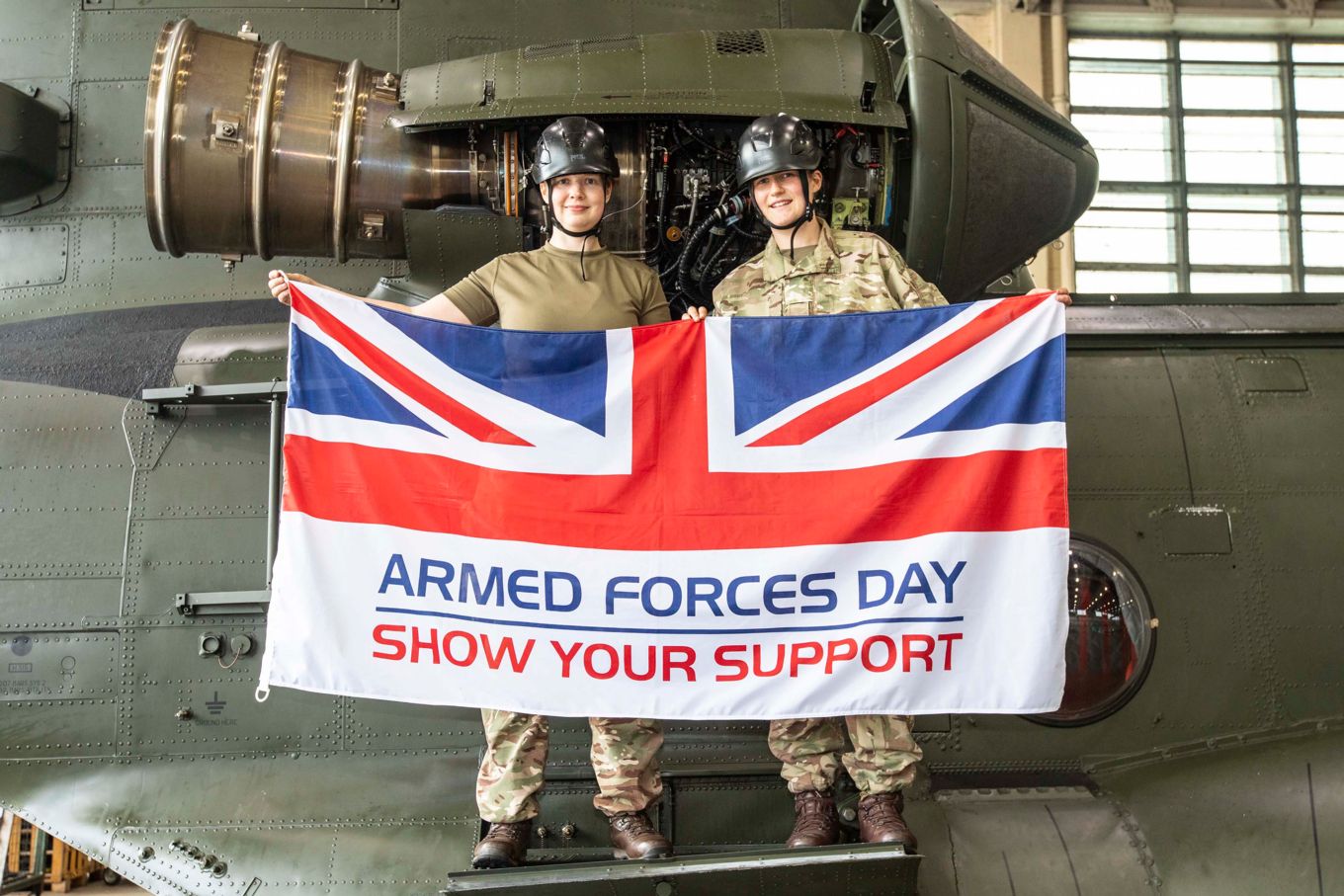 Image shows two RAF personnel holding an Armed Forces Day flag while stood on a Chinook.